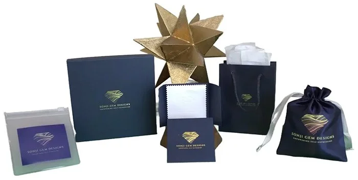 Collection of dark blue packaging with gold and white accents, including a large box, smaller boxes, a tissue box, a paper bag with tissue inside, and a drawstring pouch, all branded with Sonji Gem Designs logo, arranged around a gold geometric star sculpture.