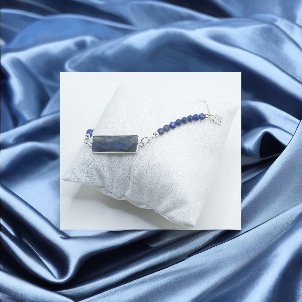 Silver bracelet with rectangular dark blue gemstone and round blue beads displayed on a white cushion, surrounded by shiny blue satin fabric.