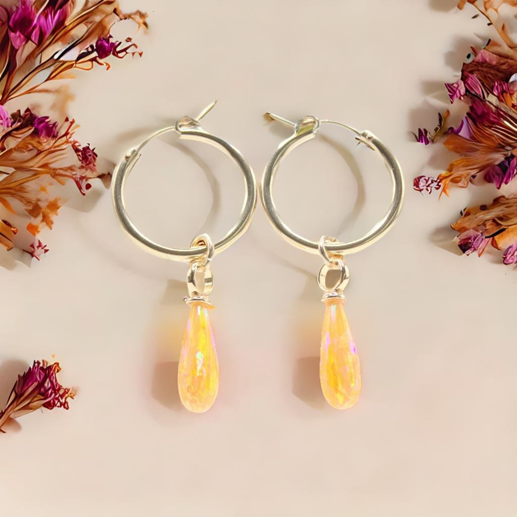 Silver hoop earrings with dangling iridescent yellow teardrop-shaped pendants, arranged on a white surface with pink and purple dried flowers around the edges.