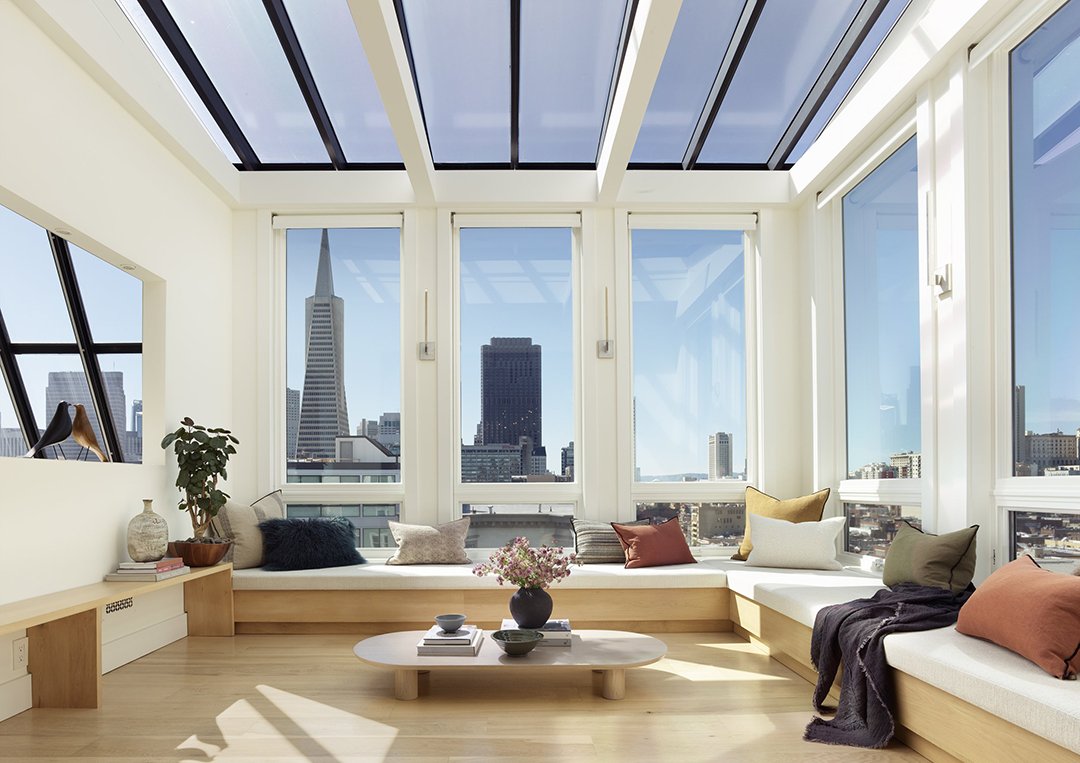 Modern sunroom with floor-to-ceiling windows and skylight, featuring a city view, built-in seating with colorful pillows, and a low coffee table with decorative items.