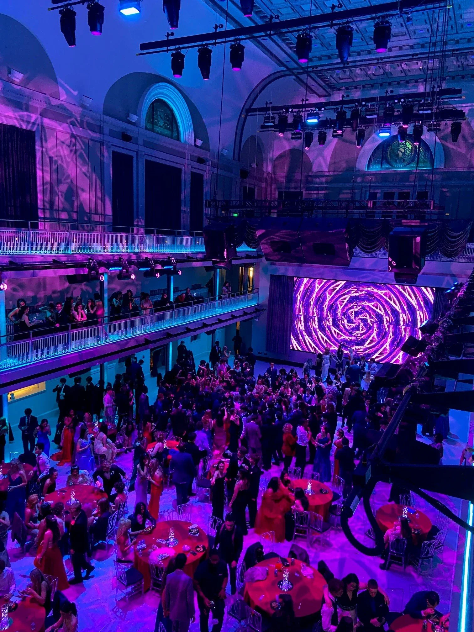 People dancing and socializing at a colorful nightclub with a large LED screen displaying purple and pink swirling visuals, multiple levels, and stage lighting.