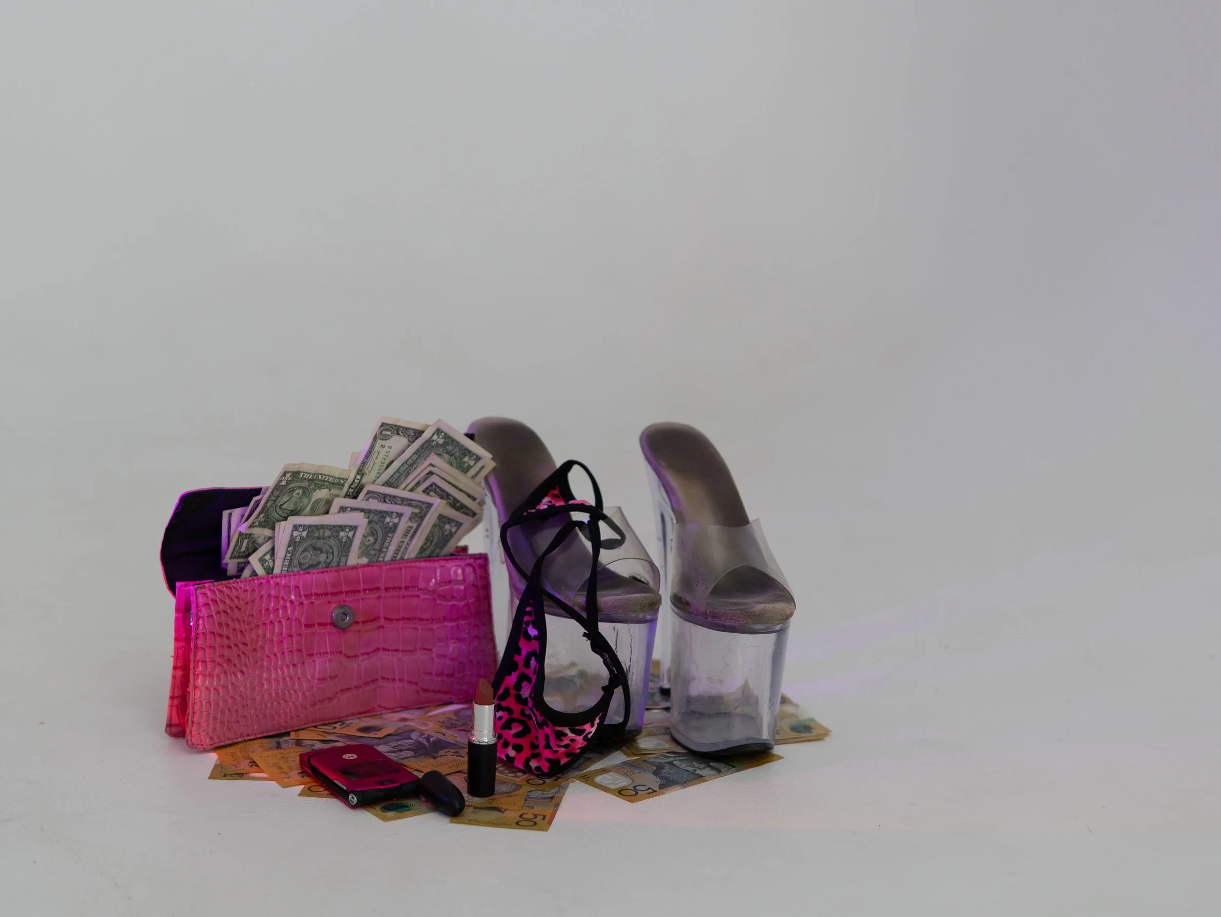Various personal items including cash, a pink purse, shoes, a lipstick, a phone, and makeup spread out on a surface with a white background.