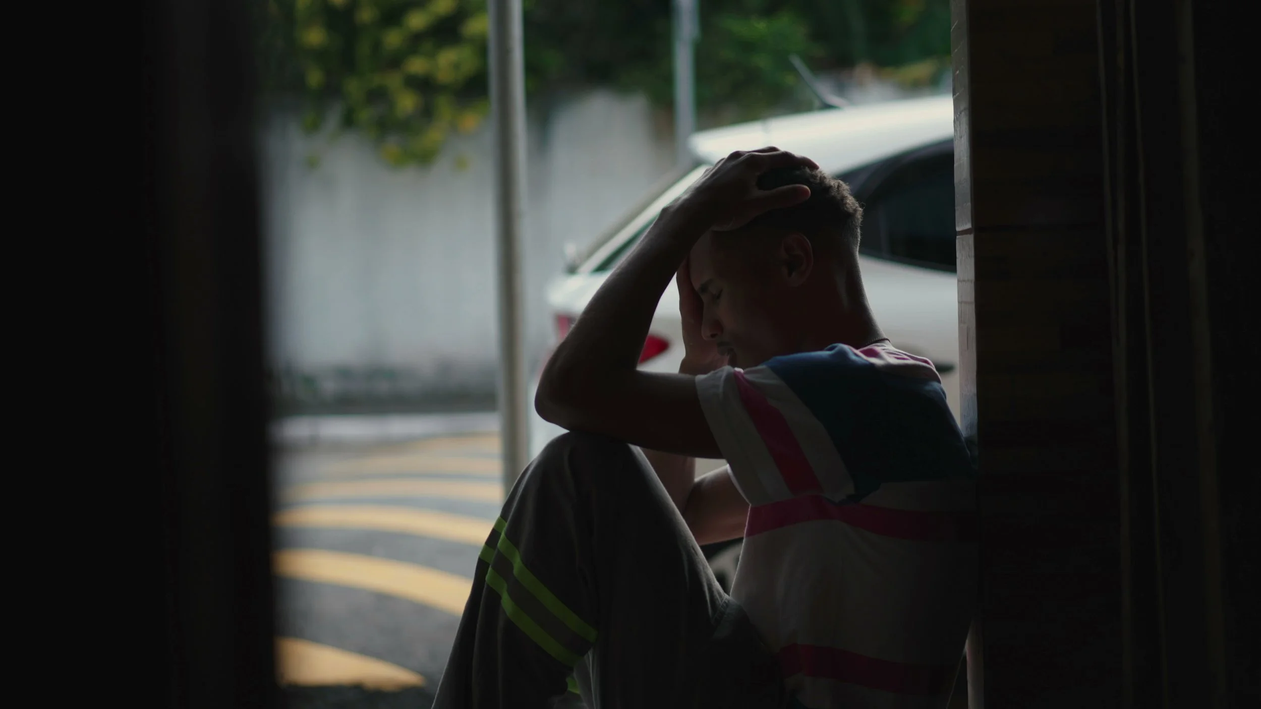 A young boy is sitting on the ground in a dark room, holding his head in his hands with a distressed expression. Outside, a car is parked on the street.