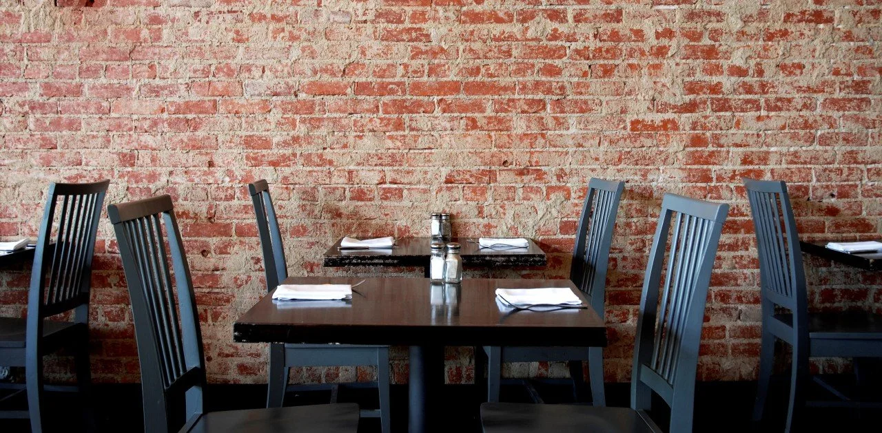 Empty restaurant table with chairs, white napkins, salt and pepper shakers against a red brick wall.