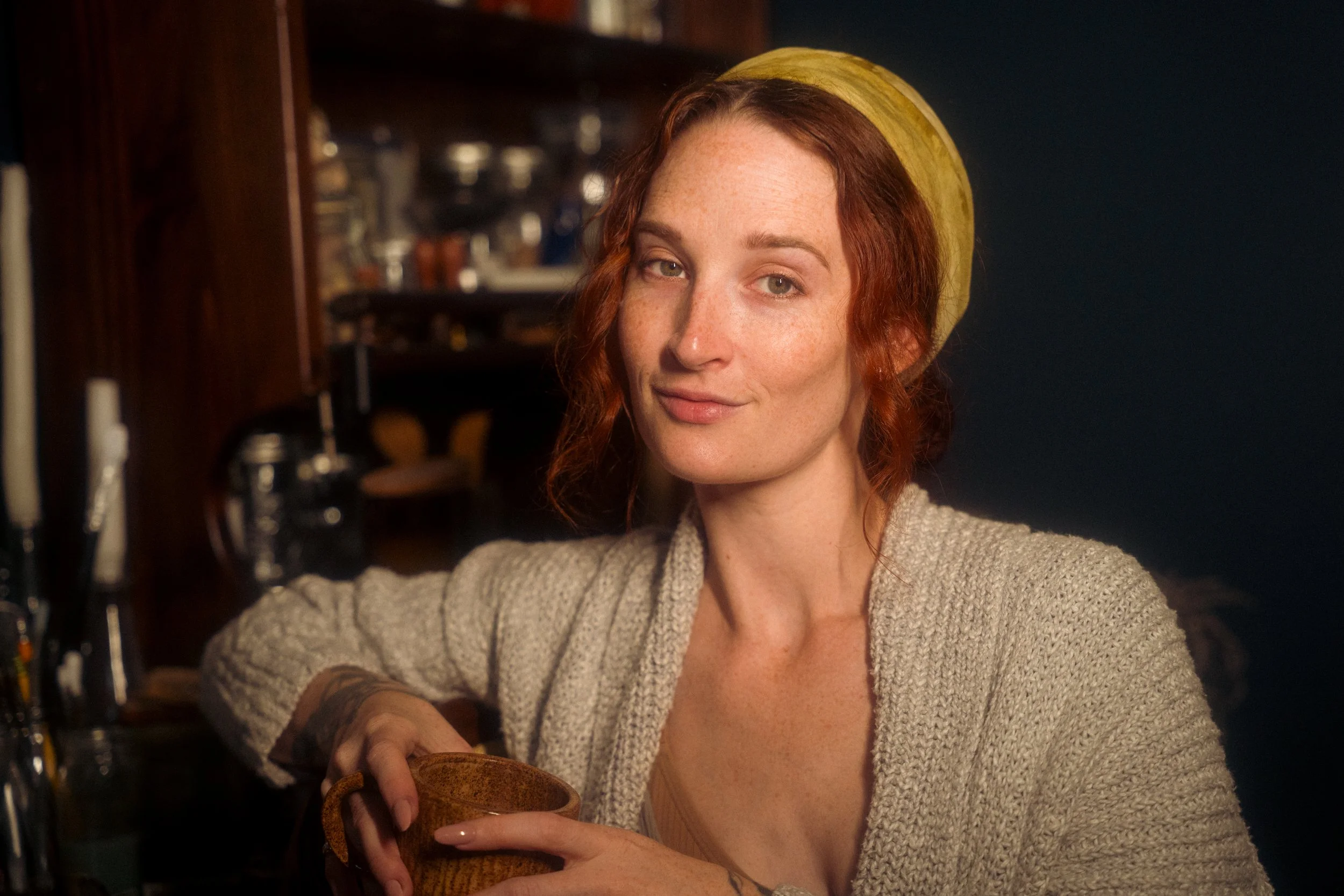 A woman with red hair, freckles, and a green headscarf sitting at a desk, holding a ceramic mug, wearing a beige sweater, with a slight smile, in a cozy indoor setting.