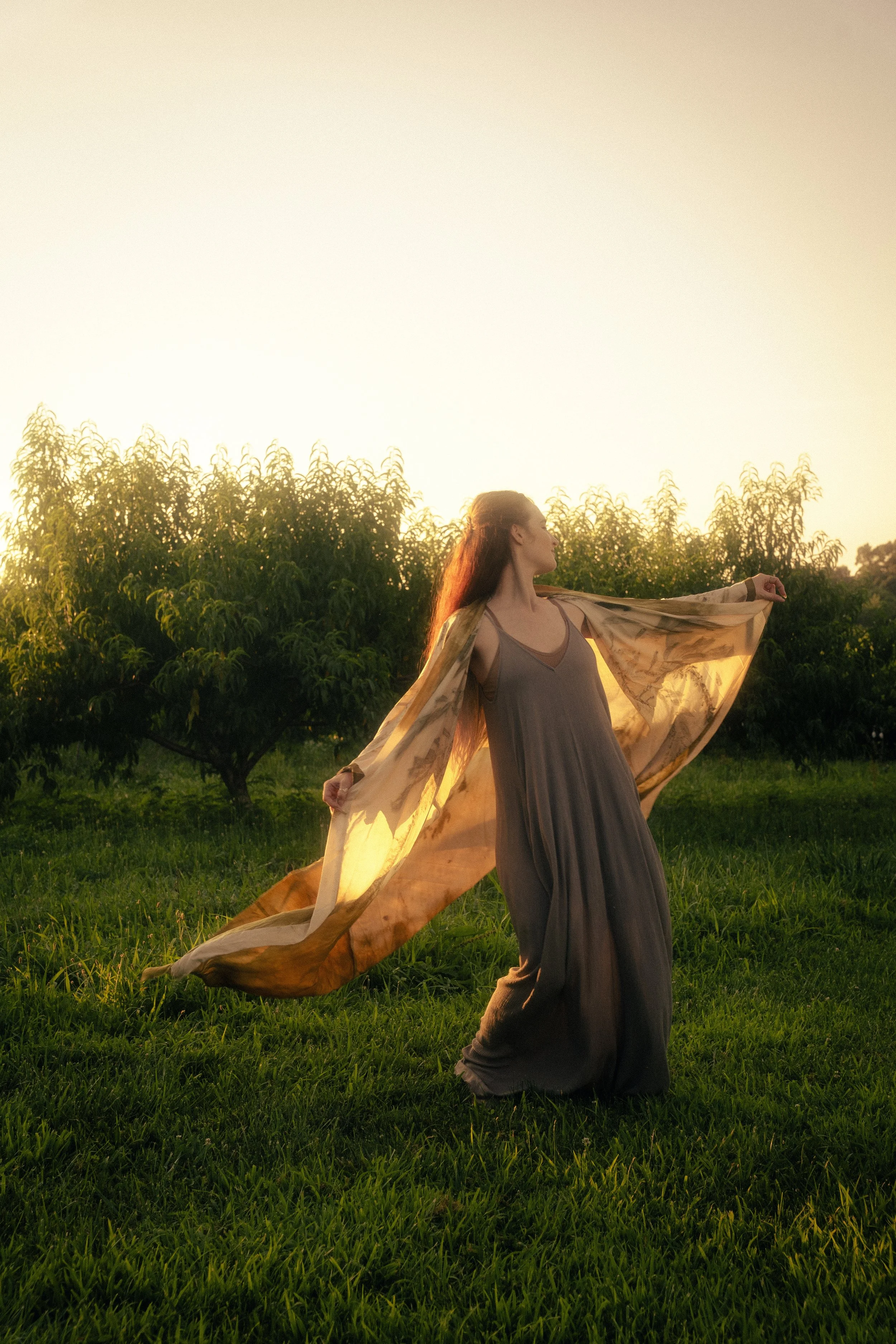 A woman standing on green grass at sunset with arms outstretched, holding a flowing robe, near trees with sunlight in the background.