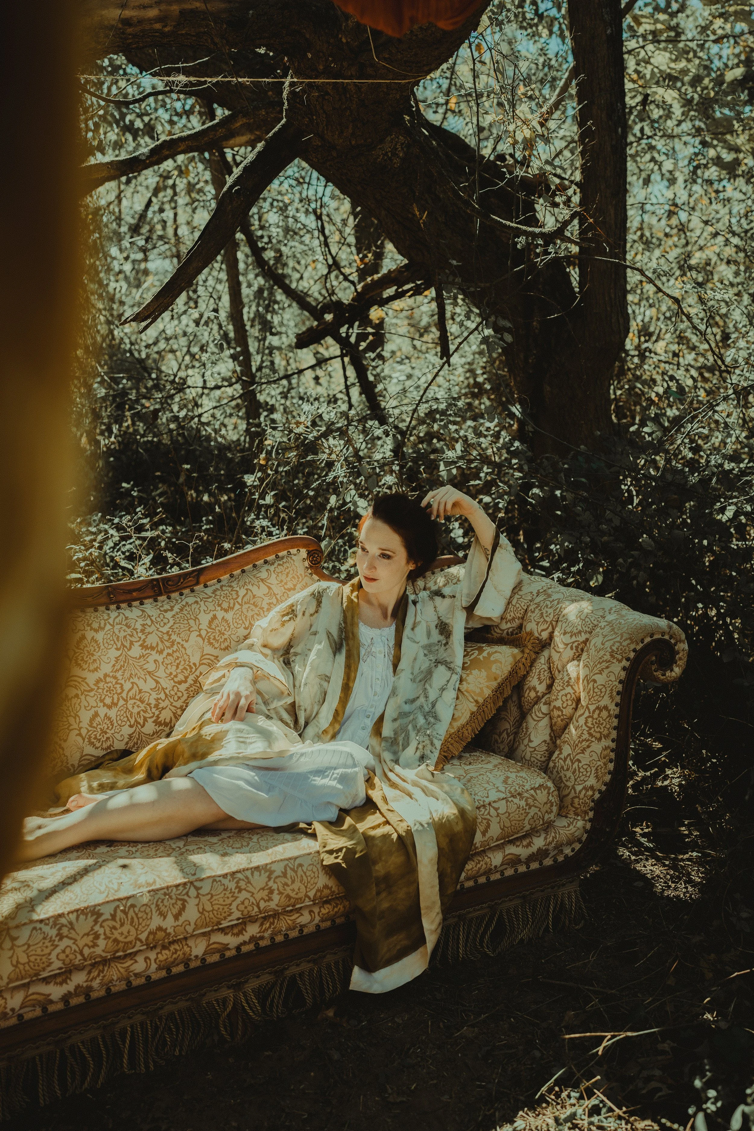 A woman dressed in vintage robes reclining on an ornate sofa outdoors in a wooded area.