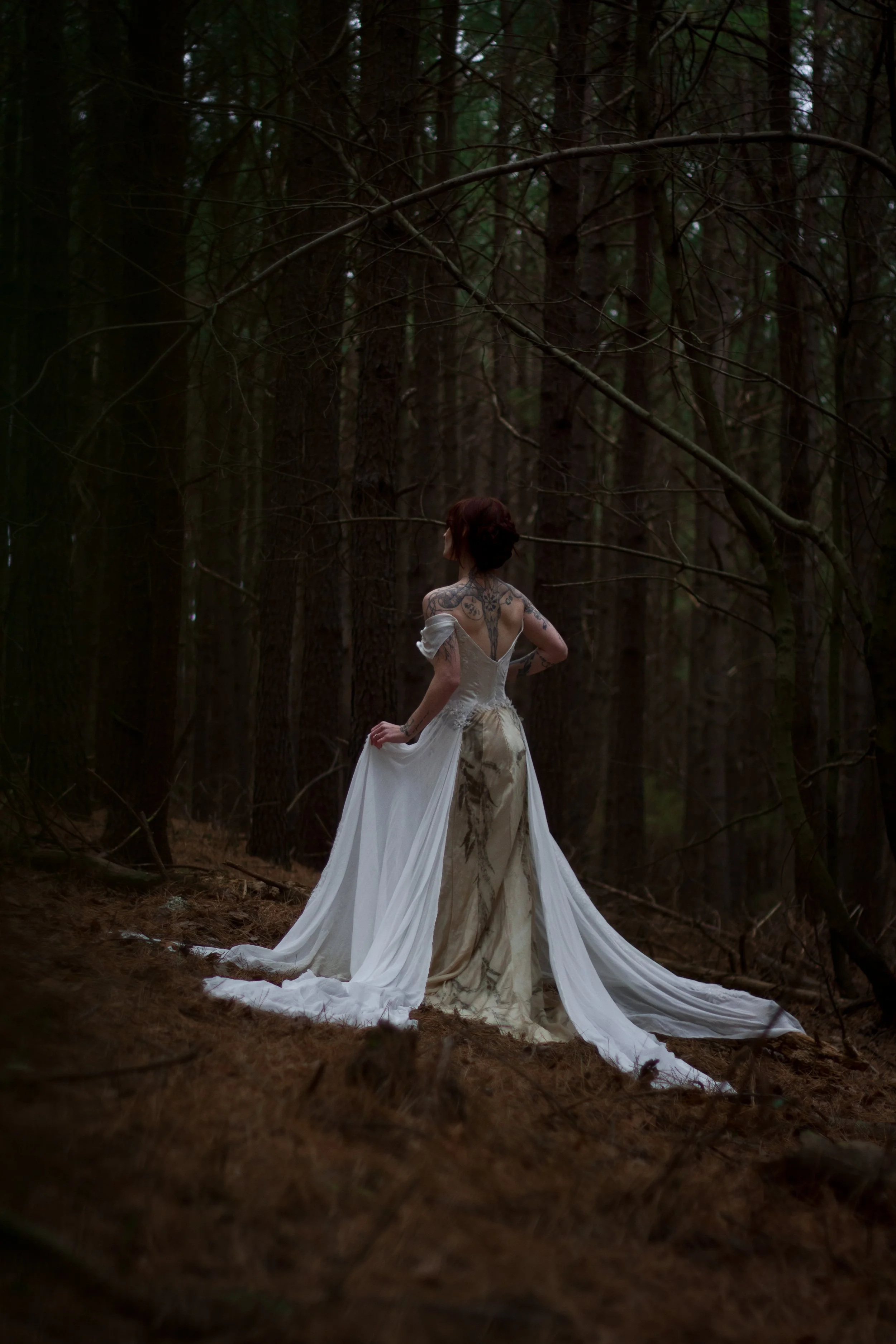 A woman with tattoos in a wedding dress standing in a dark forest.