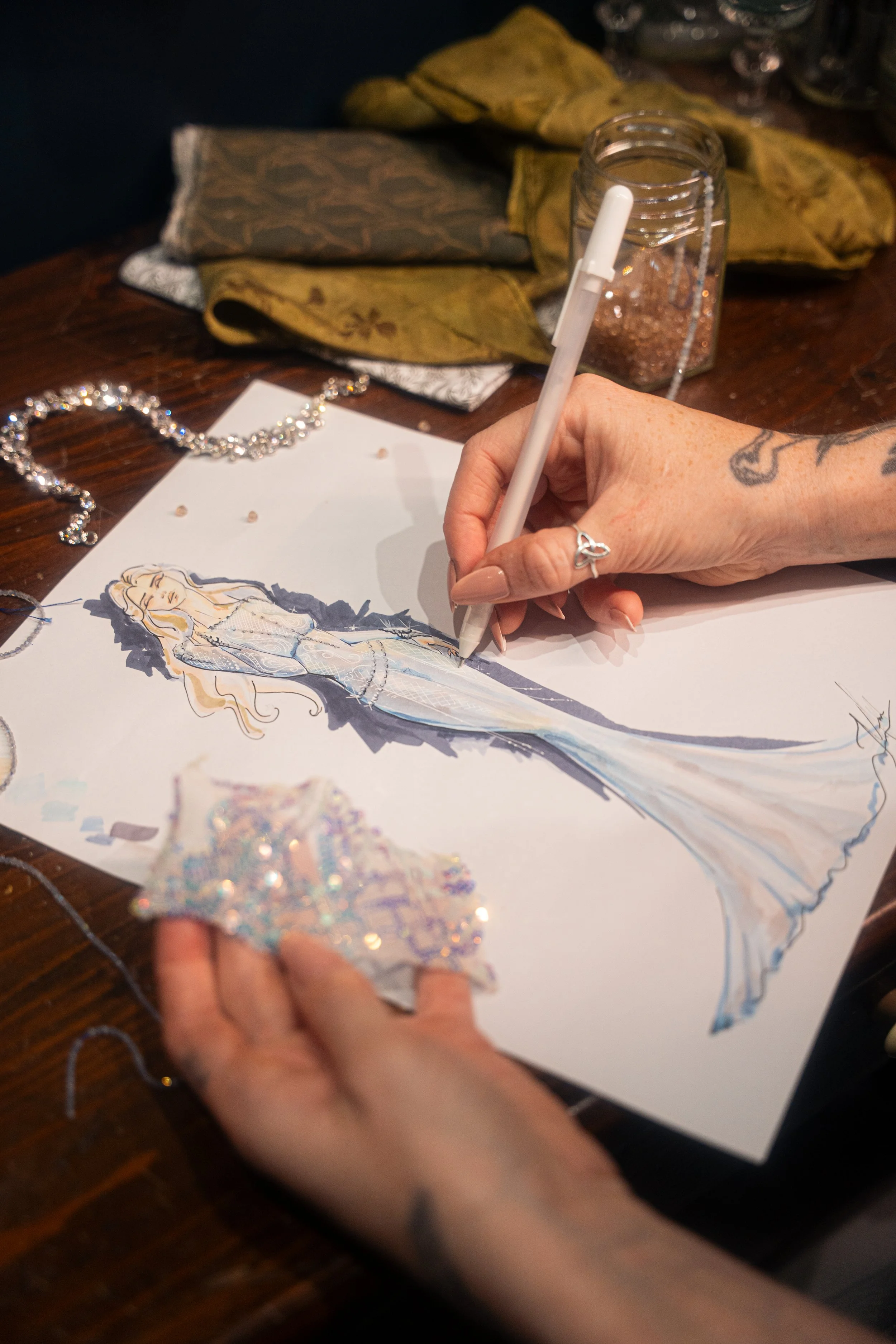An artist sketches a fashion illustration of a woman in a gown on paper, with fabric swatches and jewelry on a wooden table.