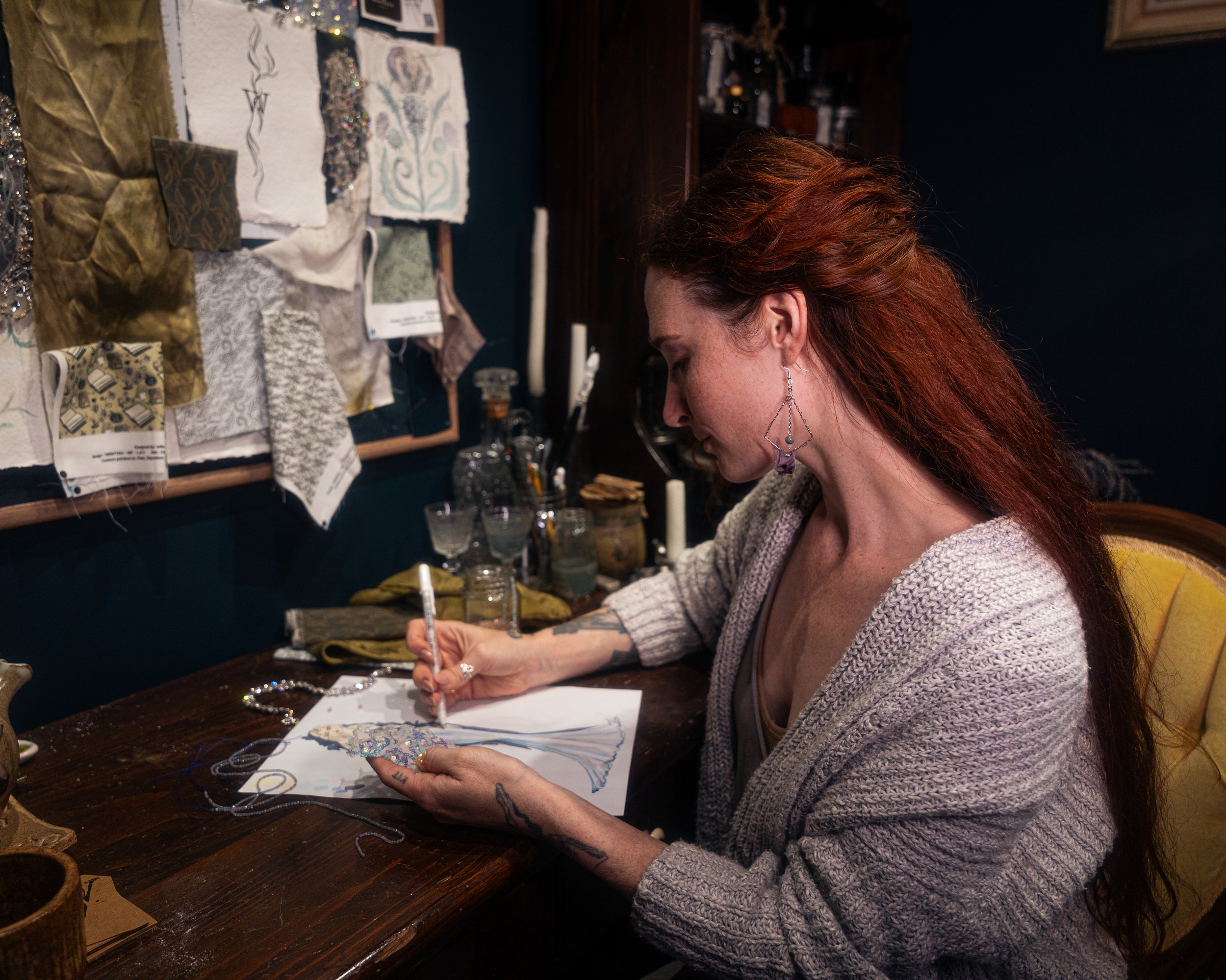 A woman with long red hair, earrings, and tattoos is sitting at a wooden table, drawing a dress on paper. The wall behind her has fabric swatches and sketches pinned up.