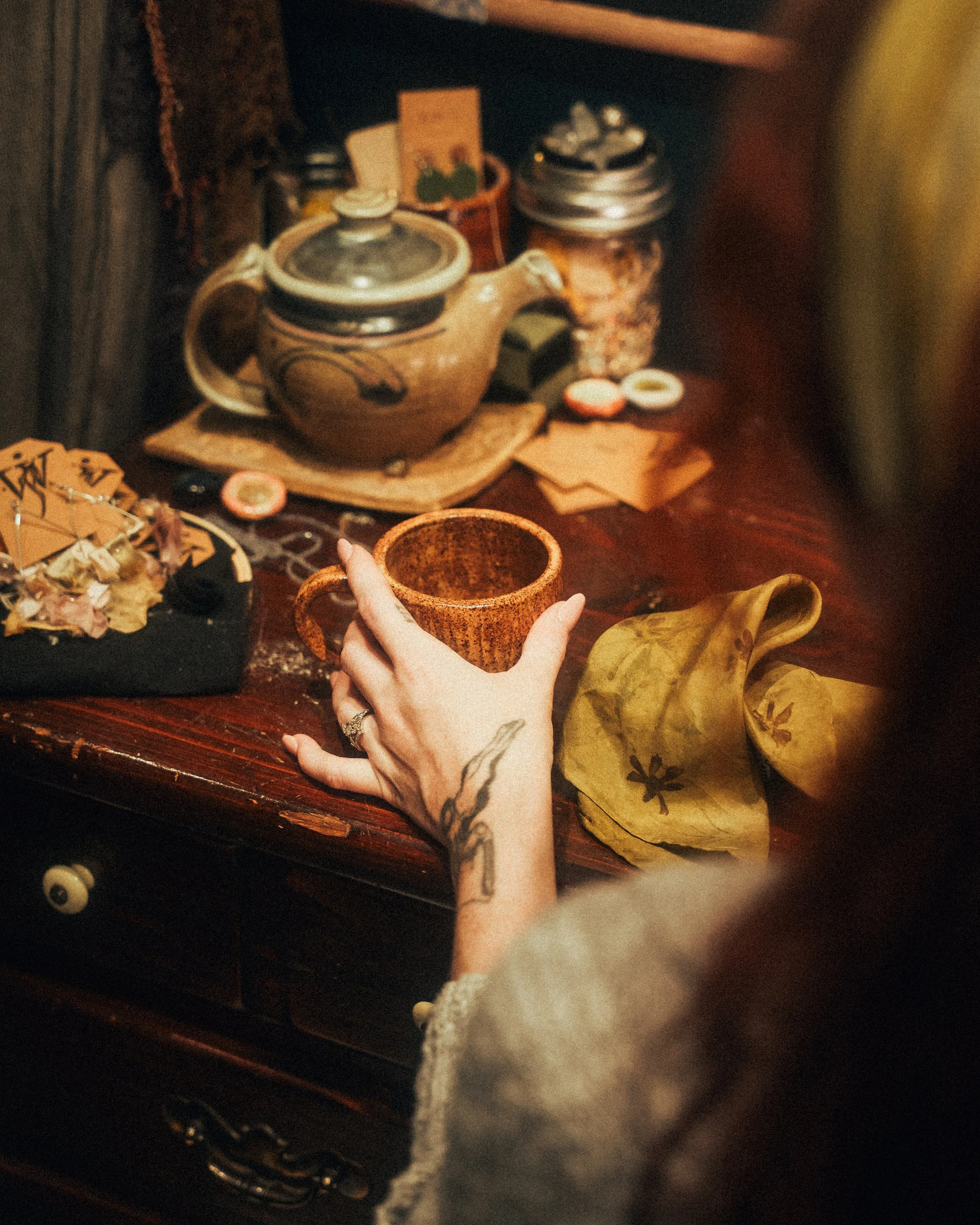 Person with tattooed arm holding a brown ceramic mug on a dark wooden table surrounded by teapots, jars, a yellow napkin, and small decorative items.