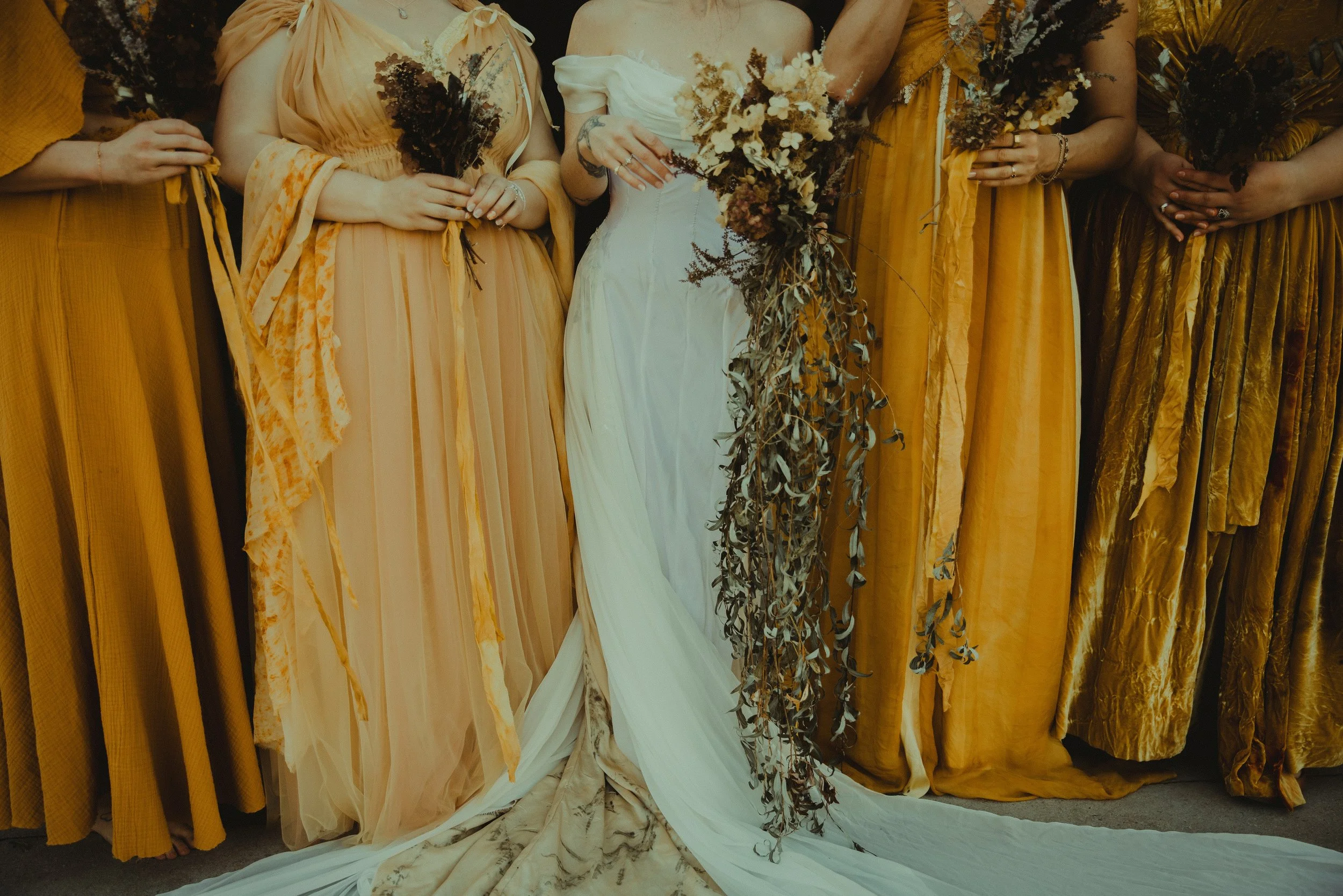 Group of women in yellow and white dresses holding bouquets of dried flowers, one woman in the center wearing a white dress with a long train.