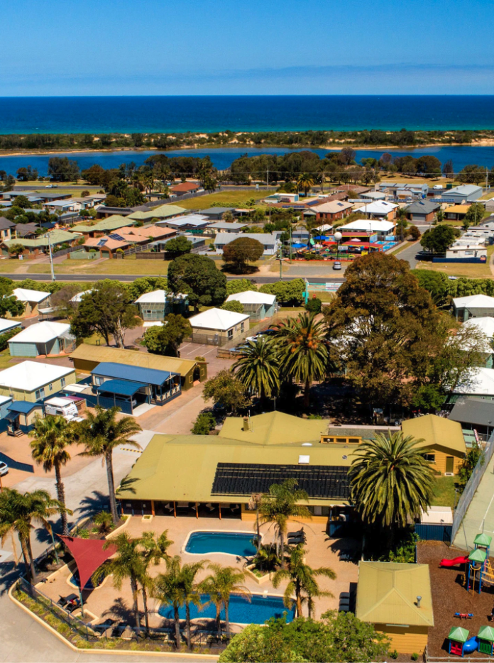 Aerial view of a coastal town with a mix of holiday homes, palm trees, and a central resort with a solar-panelled roof, near the waterfront.