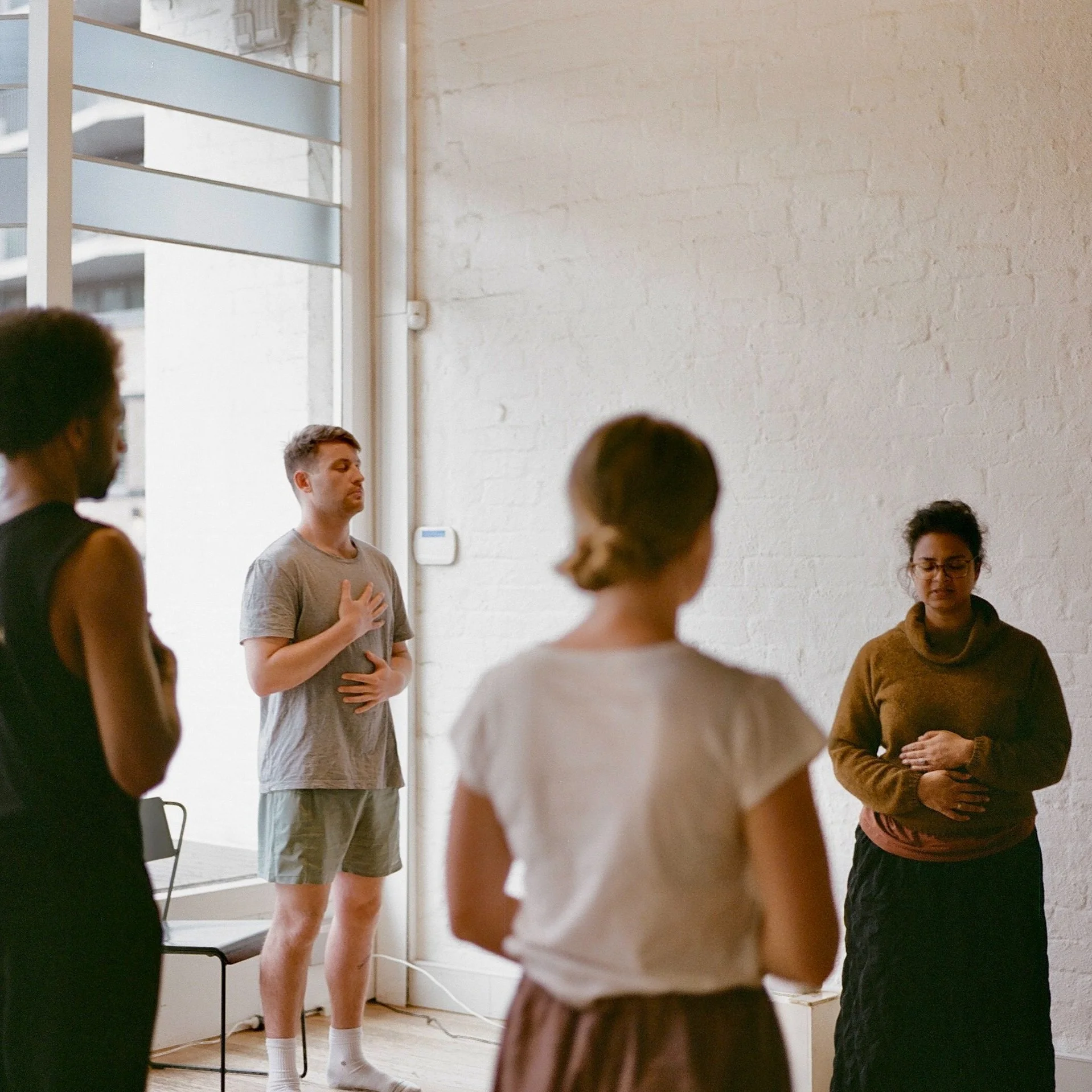 Group of four people standing in a circle with their hands on their stomachs, participating in a trauma-informed somatic mindfulness practice in a bright, airy room with white brick walls.