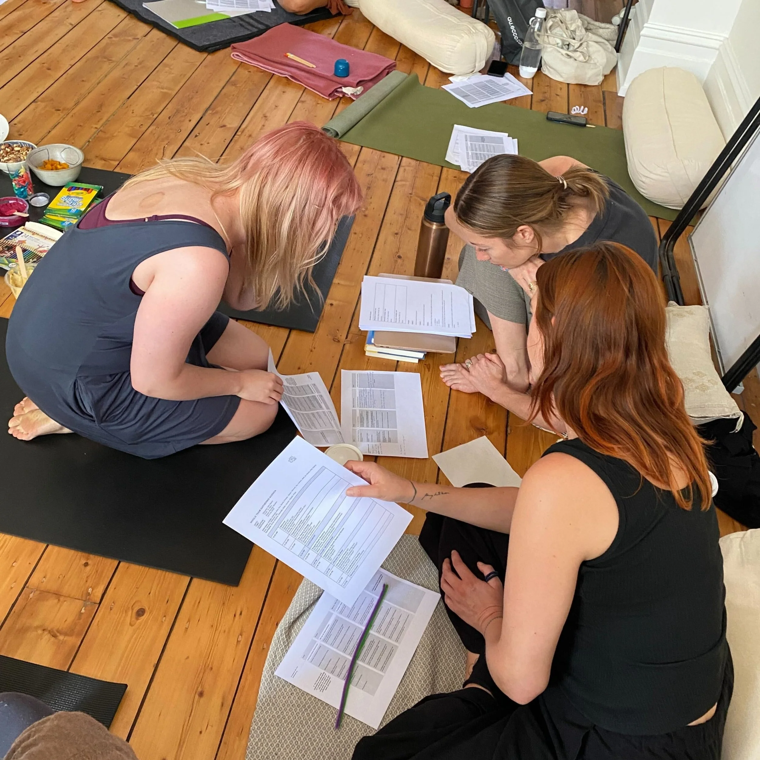 Three women sitting and kneeling on yoga mats on a wooden floor, reviewing papers and documents together in a casual indoor setting.