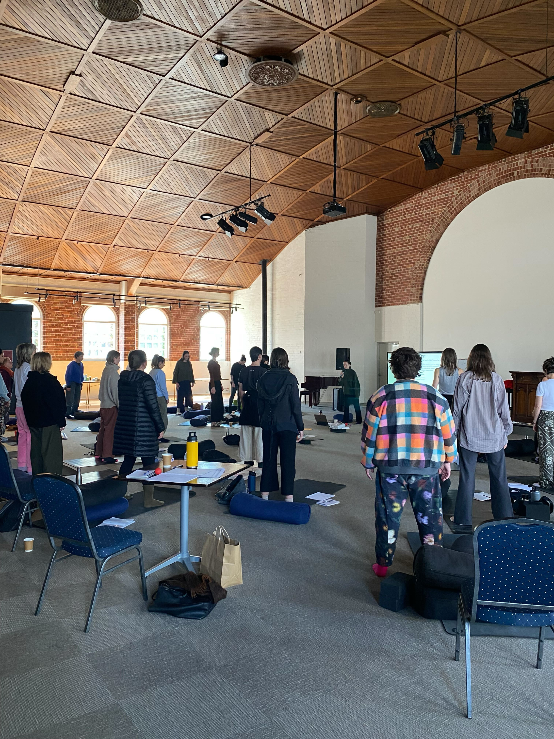 A group of people standing in a spacious room with wooden ceiling panels, at a somatic therapy training.There are mats on the floor, and some personal belongings such as water bottles, bags, and notebooks. The room has large arched windows. 