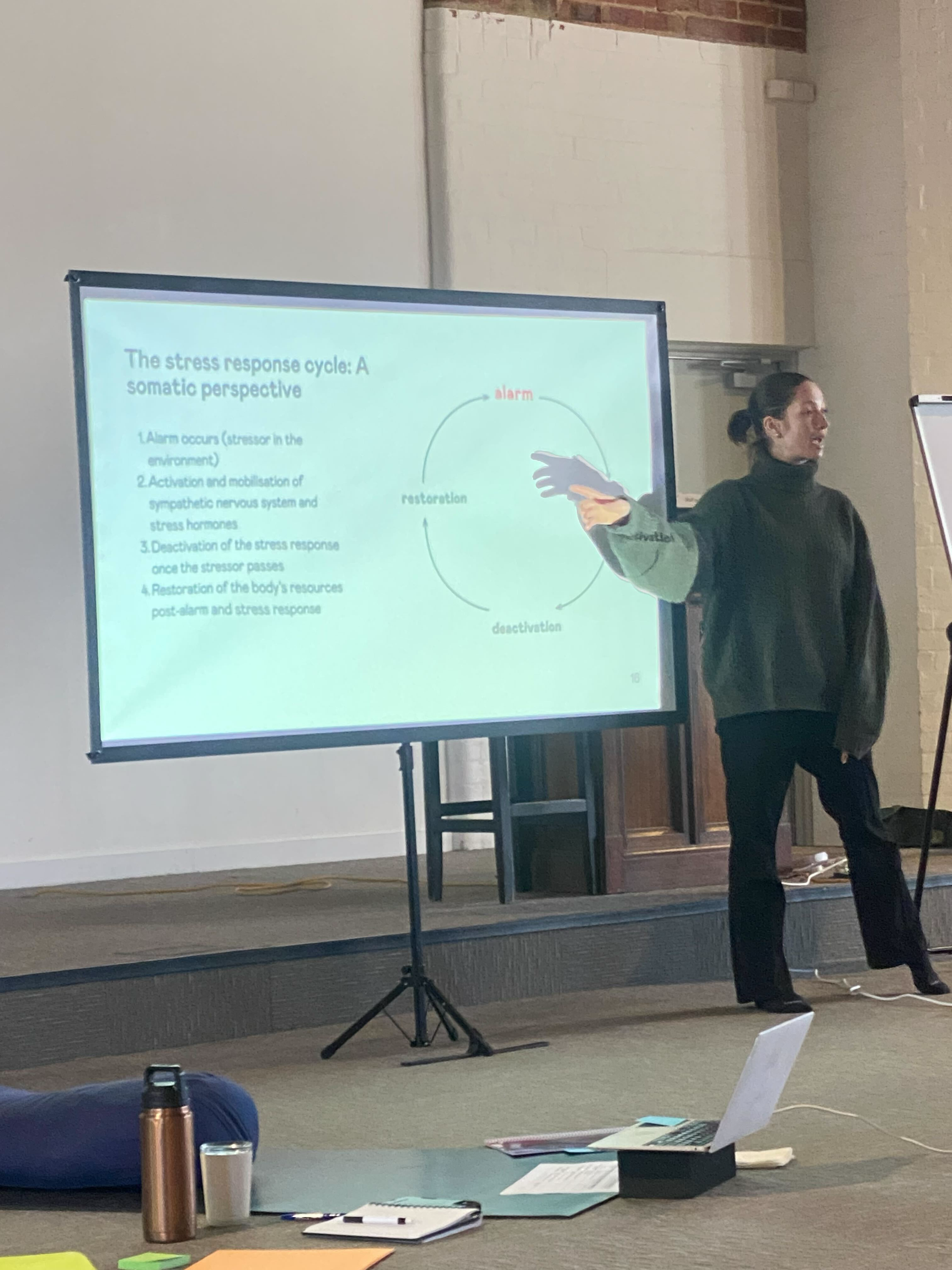 A woman giving a presentation on the stress response cycle at a trauma-informed, somatic training, standing next to a screen displaying a diagram and text.