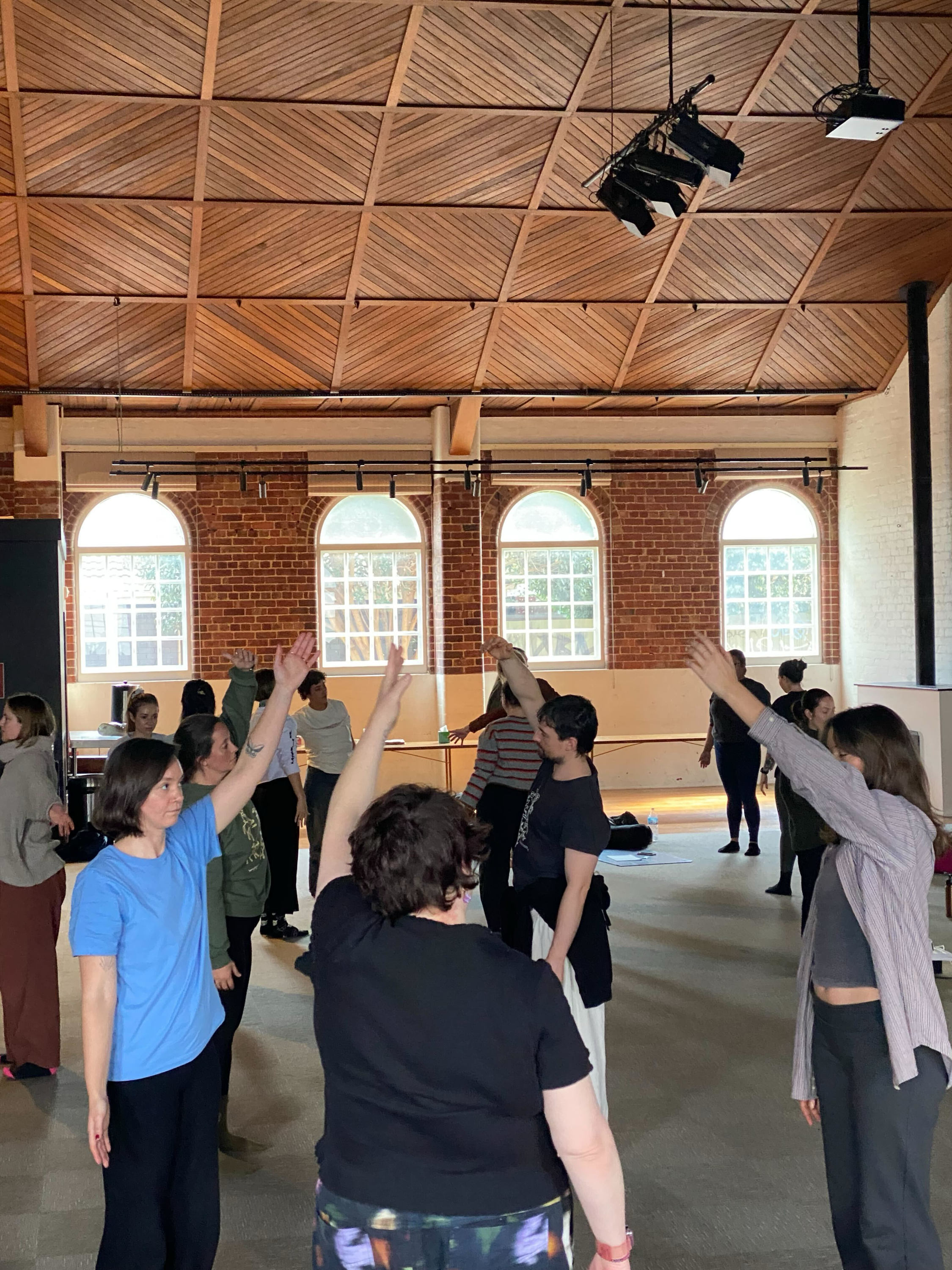 Group of people participating in a somatic training in Melbourne, raising their hands, under a wooden ceiling with brick walls and large arched windows.