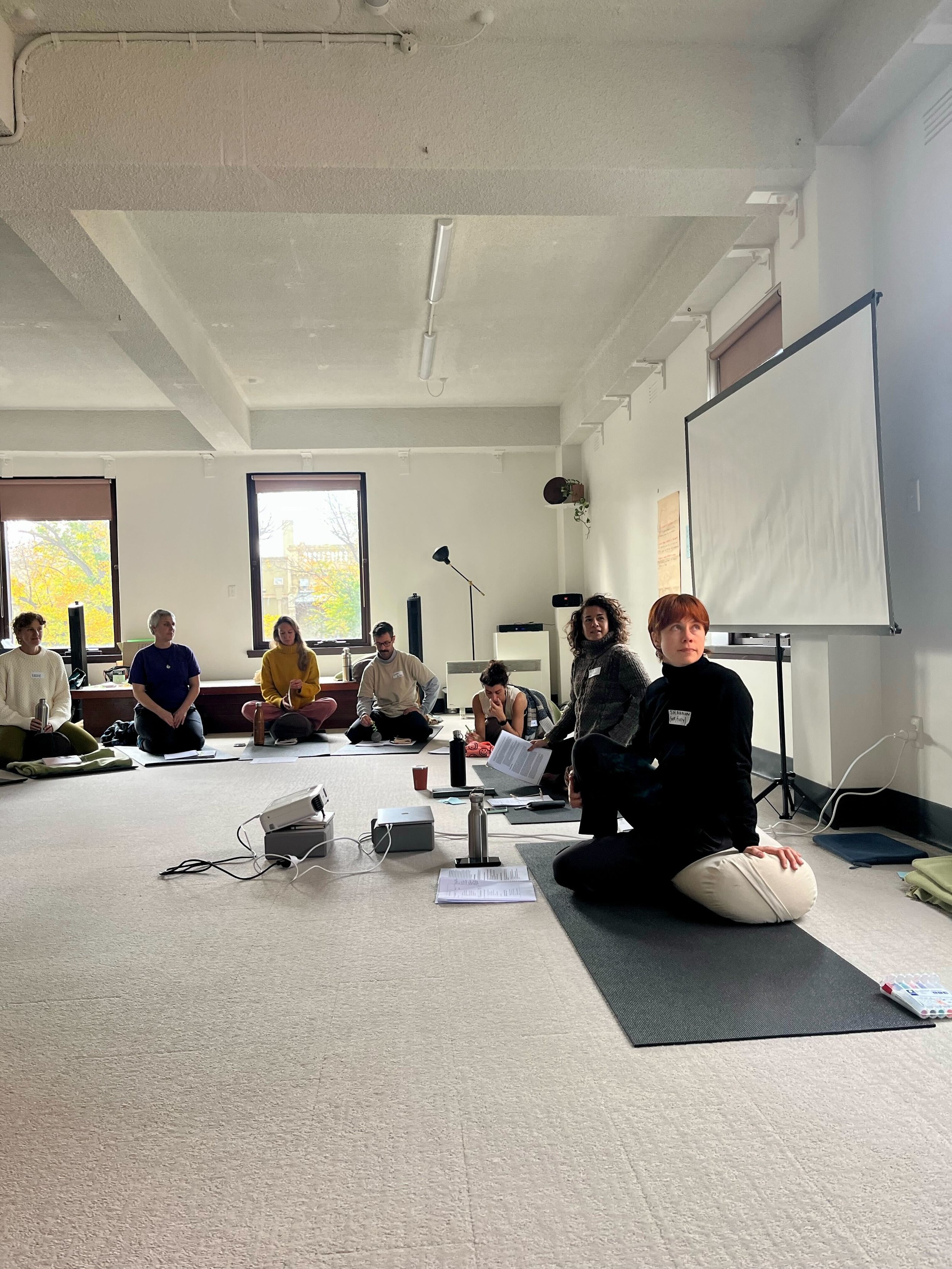 Group of people sitting on the floor in a circle during a trauma-informed somatic training workshop in a bright room with large windows, some with laptops, notebooks, and water bottles.
