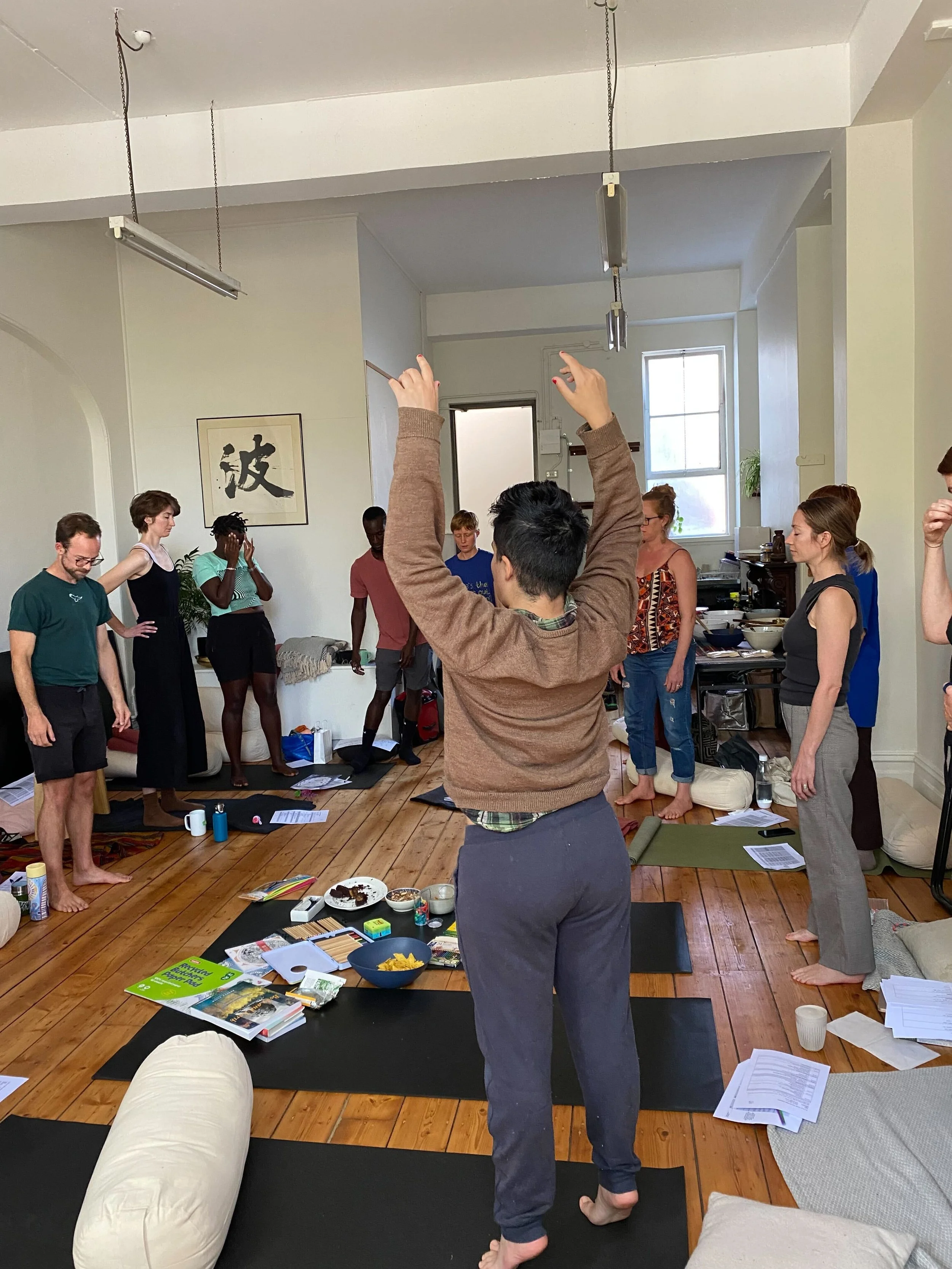 A group of people in a room participating in a somatics training, with one person standing with arms raised, facing away from the camera, and others standing or sitting on yoga mats