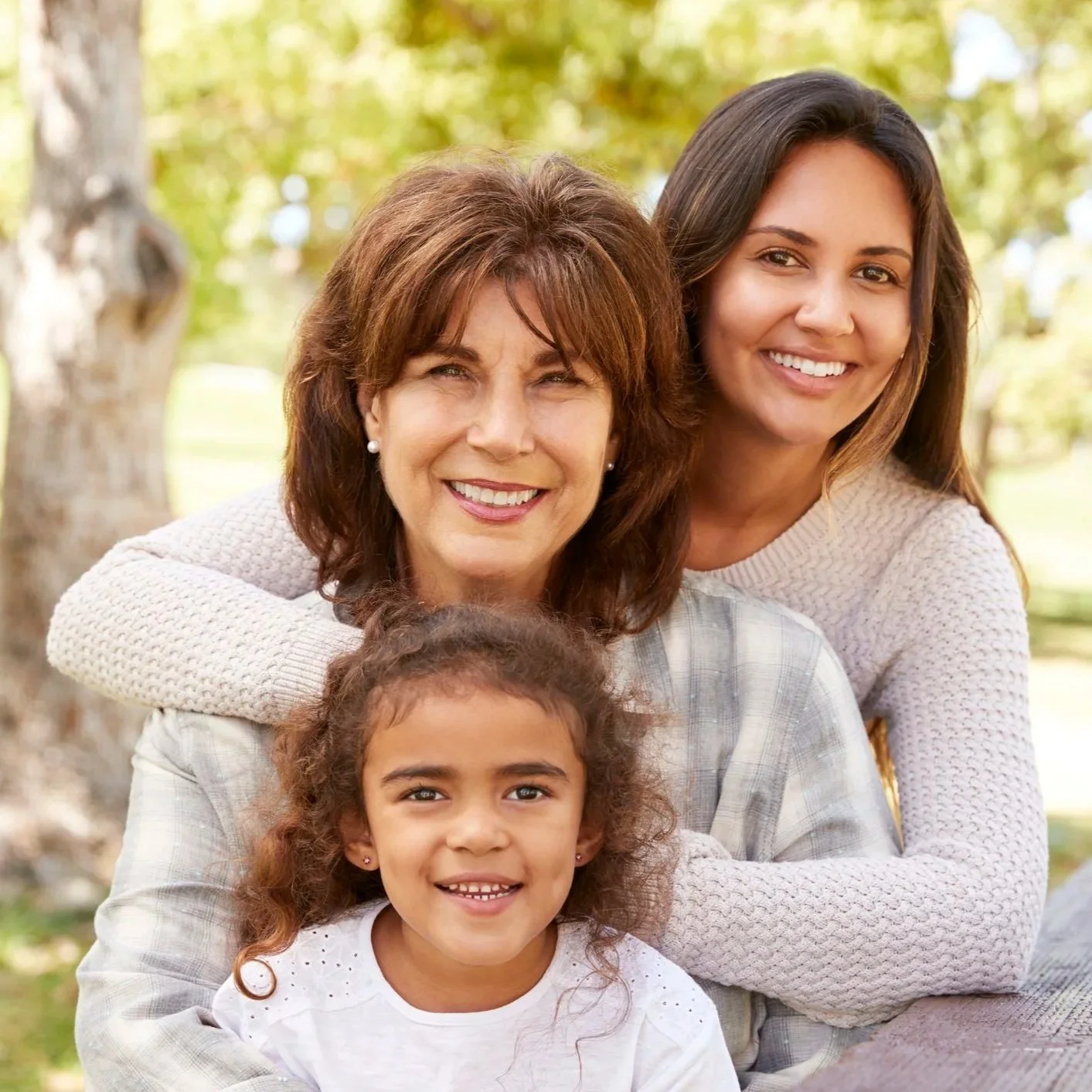 Three generations of women smiling outdoors in a park with autumn trees in background