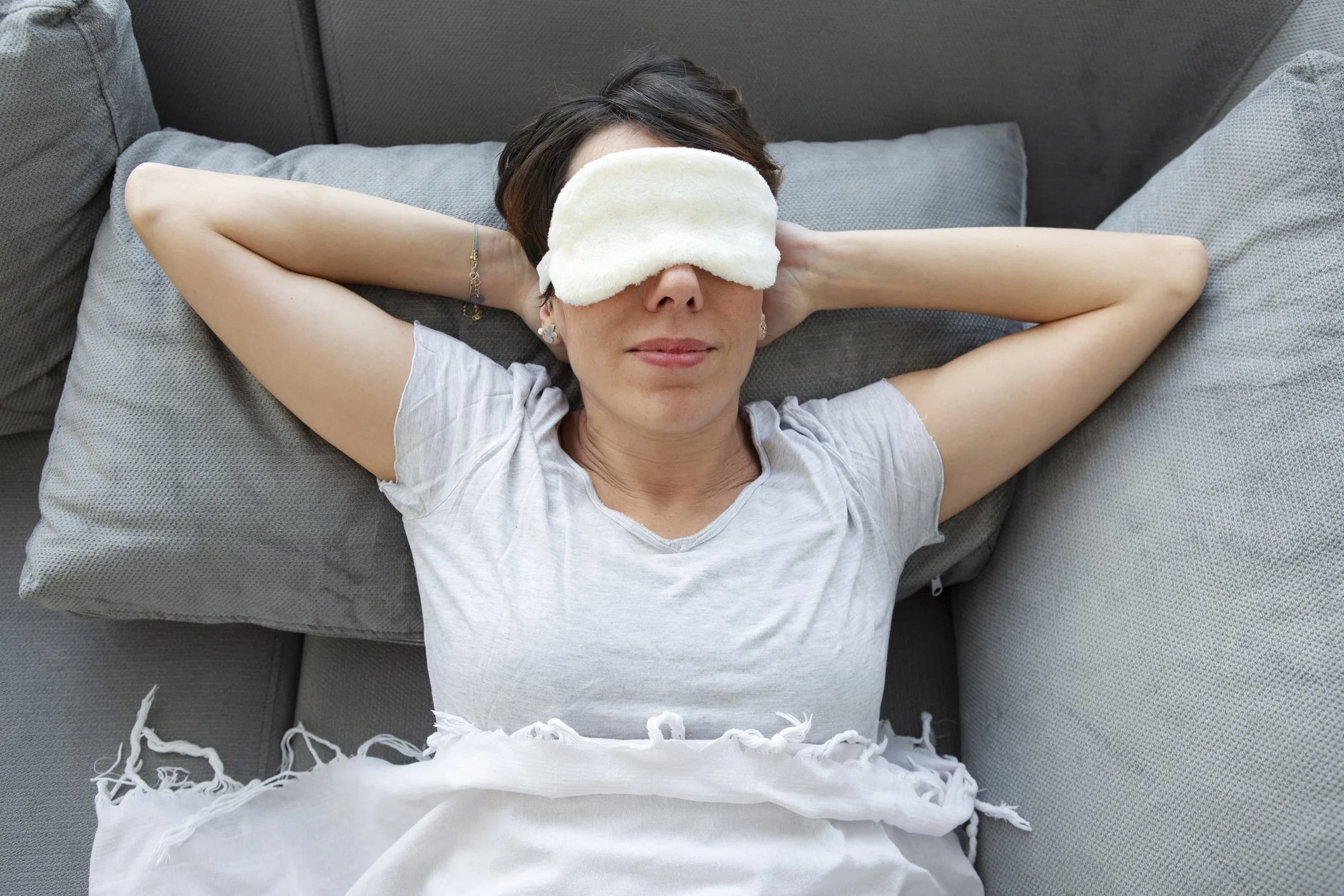 A woman lying on a grey sofa with her eyes closed, wearing a sleep mask, a grey t-shirt, and surrounded by grey pillows.