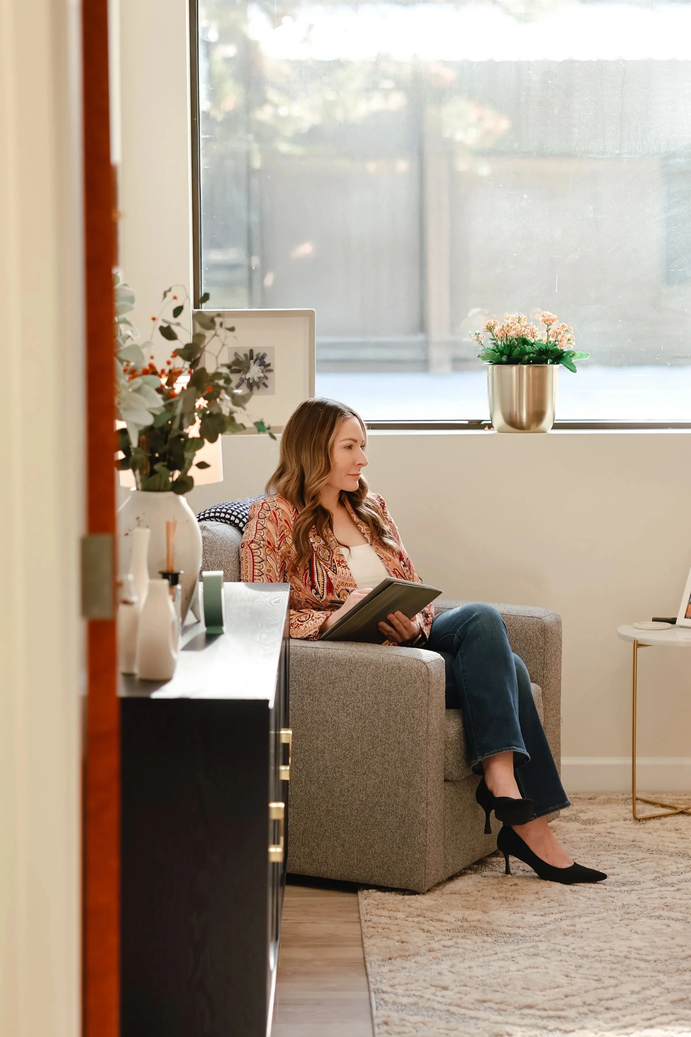A woman therapist sitting on a gray sofa in a bright room, holding a notebook, with a large window and potted plant behind her.