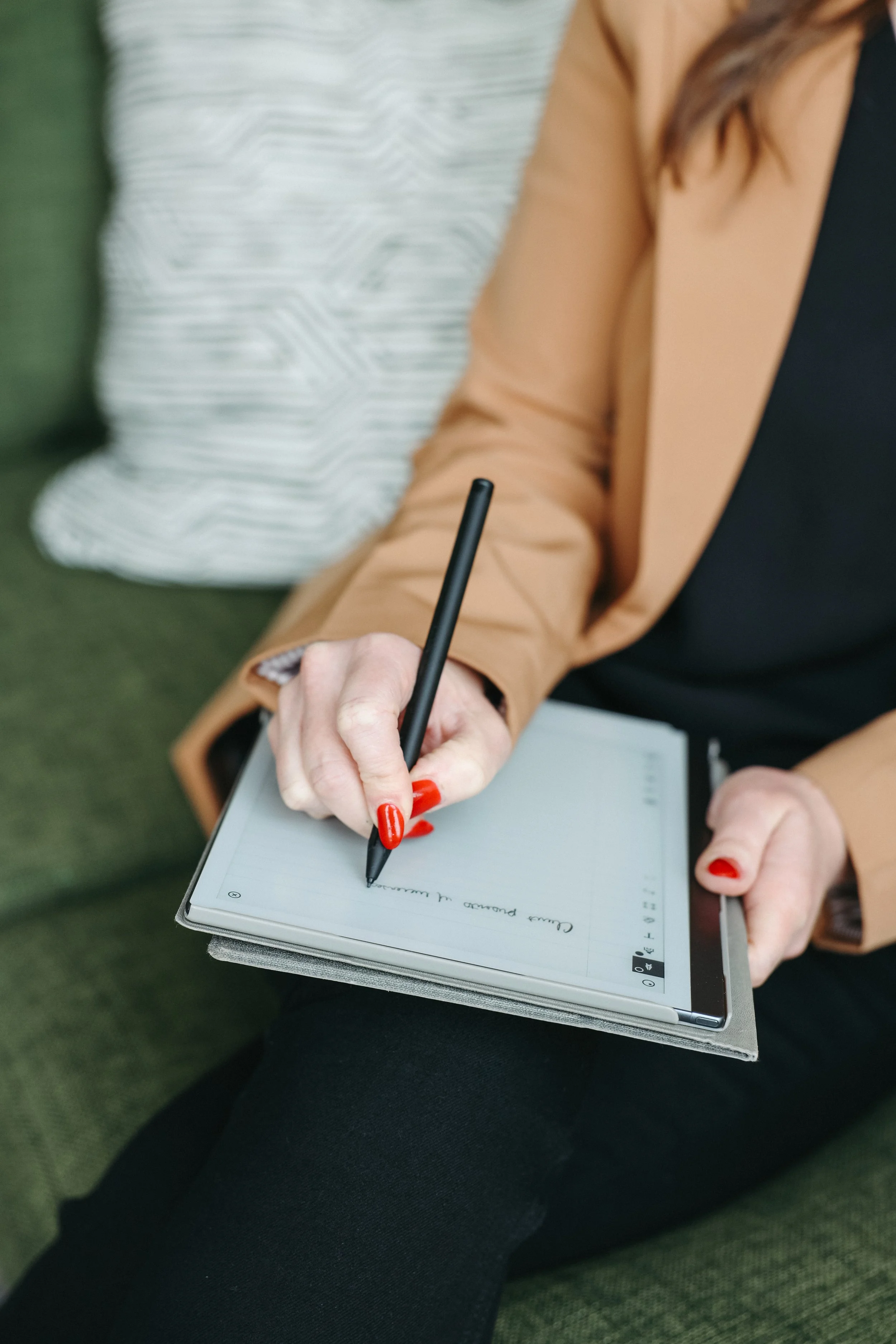 A woman with red painted nails writes in a digital notebook with a stylus, sitting on a green couch.