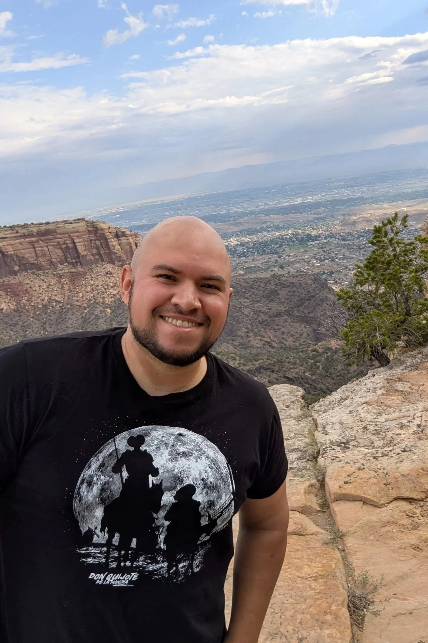 A smiling man with a beard and a black T-shirt featuring a moon and silhouette of Don Quixote and Sancho Panza, standing on rocks with a canyon landscape and cloudy sky in the background.