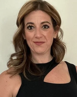 A woman with wavy, light brown hair and a black sleeveless top, smiling slightly at the camera against a plain background.