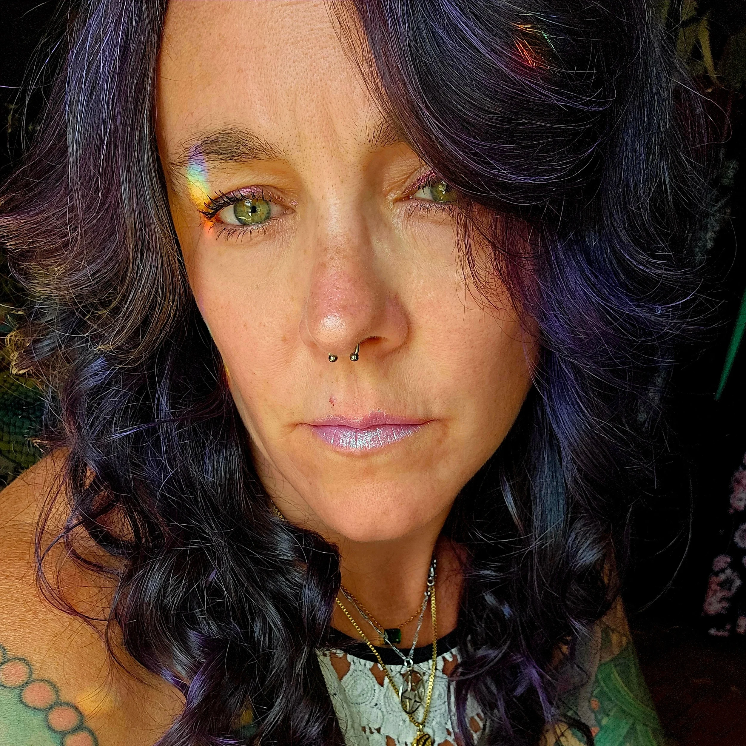 A woman with dark, wavy hair and light green eyes, wearing jewelry including necklaces and septum piercing. She has makeup with shimmery lipstick and slight eye makeup, and her hair has hints of purple.