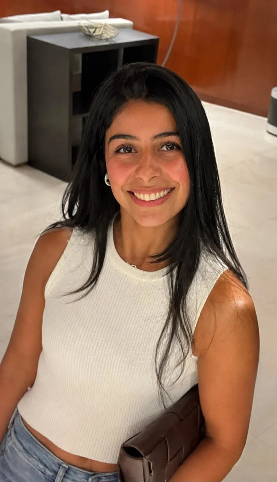 A woman with black hair, smiling and wearing a white sleeveless top, sitting in an indoor setting.