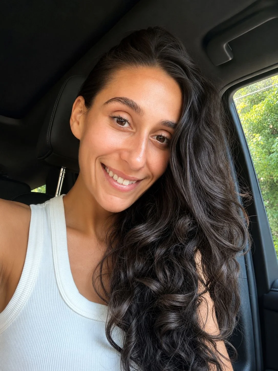 A smiling woman with long, curly dark hair sitting in a car, with green trees visible through the window.