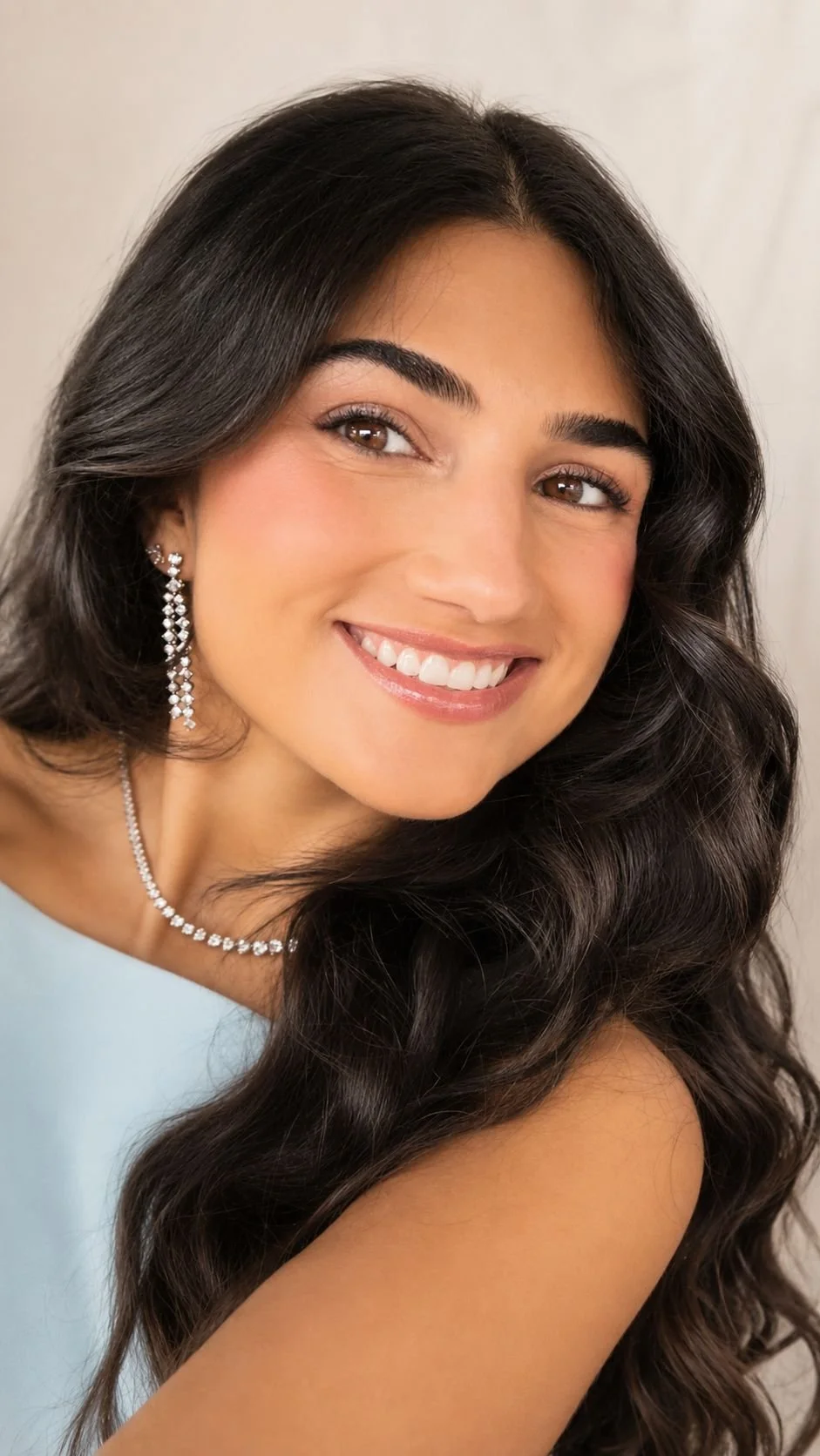 Close-up of a smiling woman with long dark hair, wearing diamond jewelry and a light-colored top.