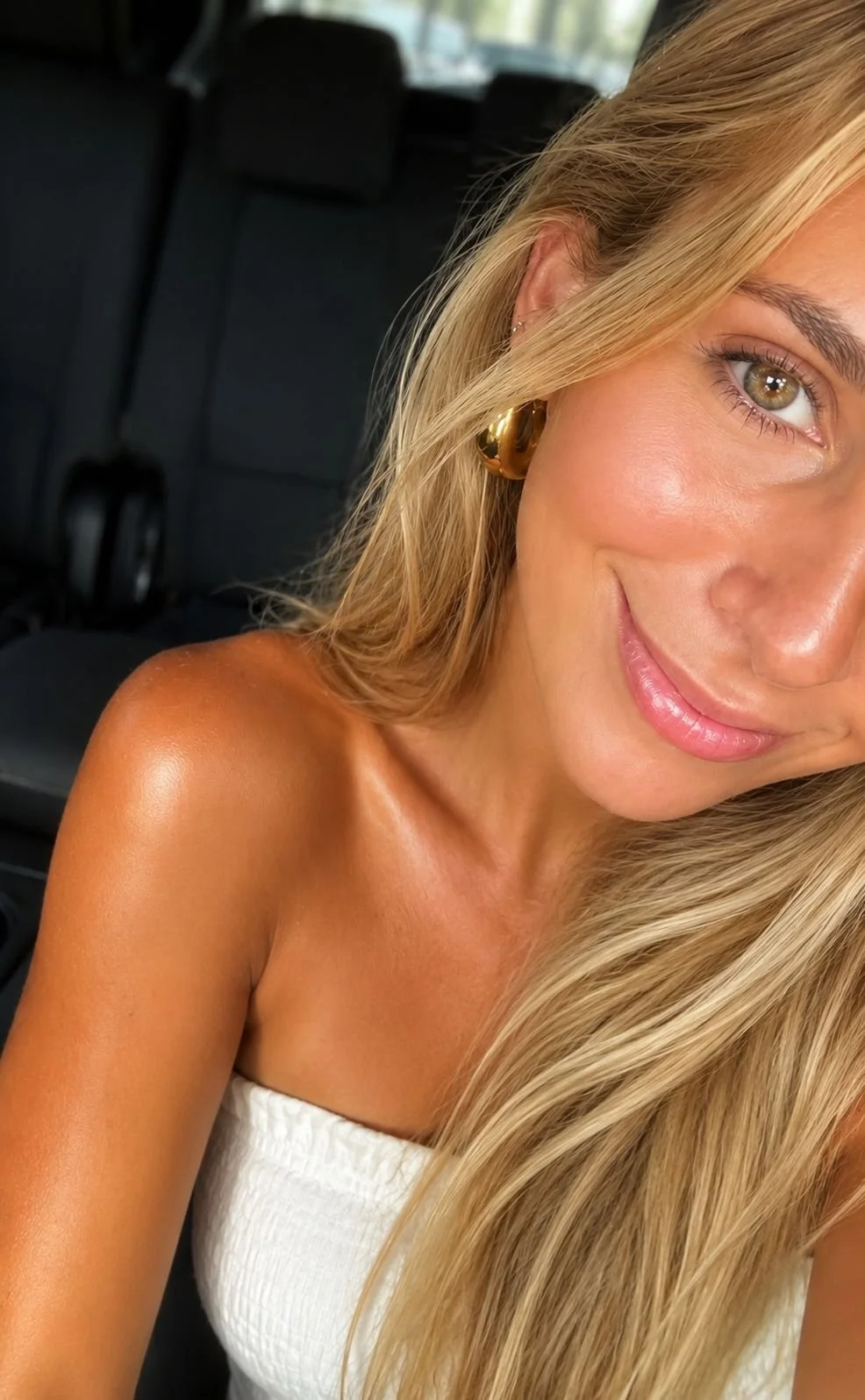 Close-up photo of a smiling woman with long blonde hair, wearing gold hoop earrings, inside a vehicle.