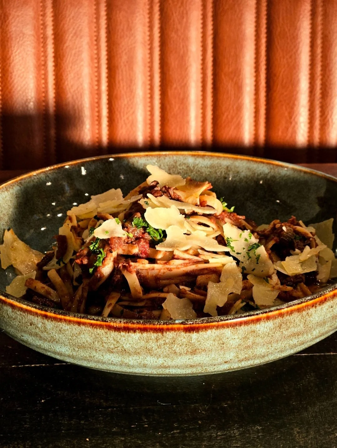 A bowl of pasta with shaved cheese, herbs, and possibly mushrooms, on a dark wooden table with a textured curtain in the background.