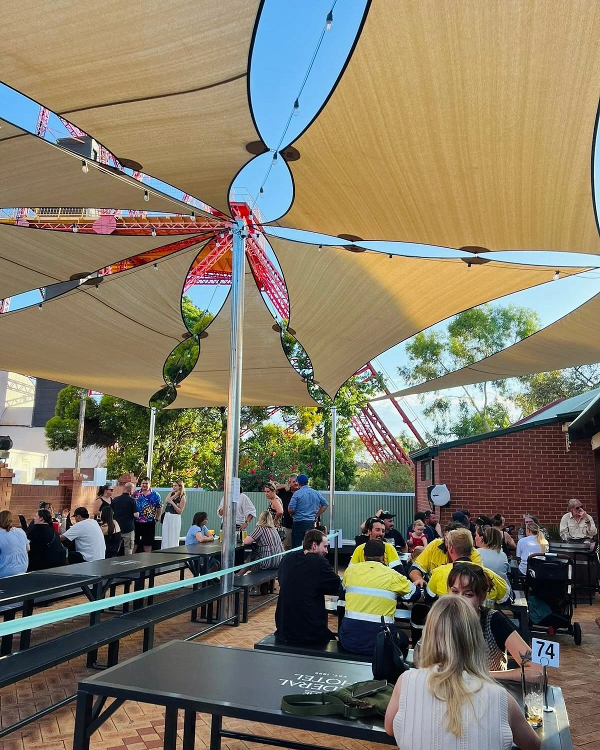People dining and socializing under shade sails at an outdoor venue, with a roller coaster visible in the background.