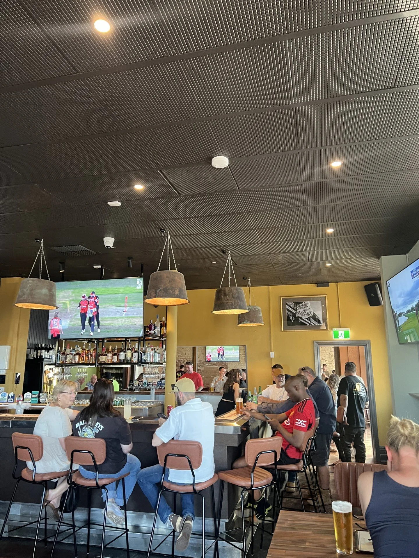 People sitting and standing at a bar in a sports bar, watching multiple screens showing a football game.