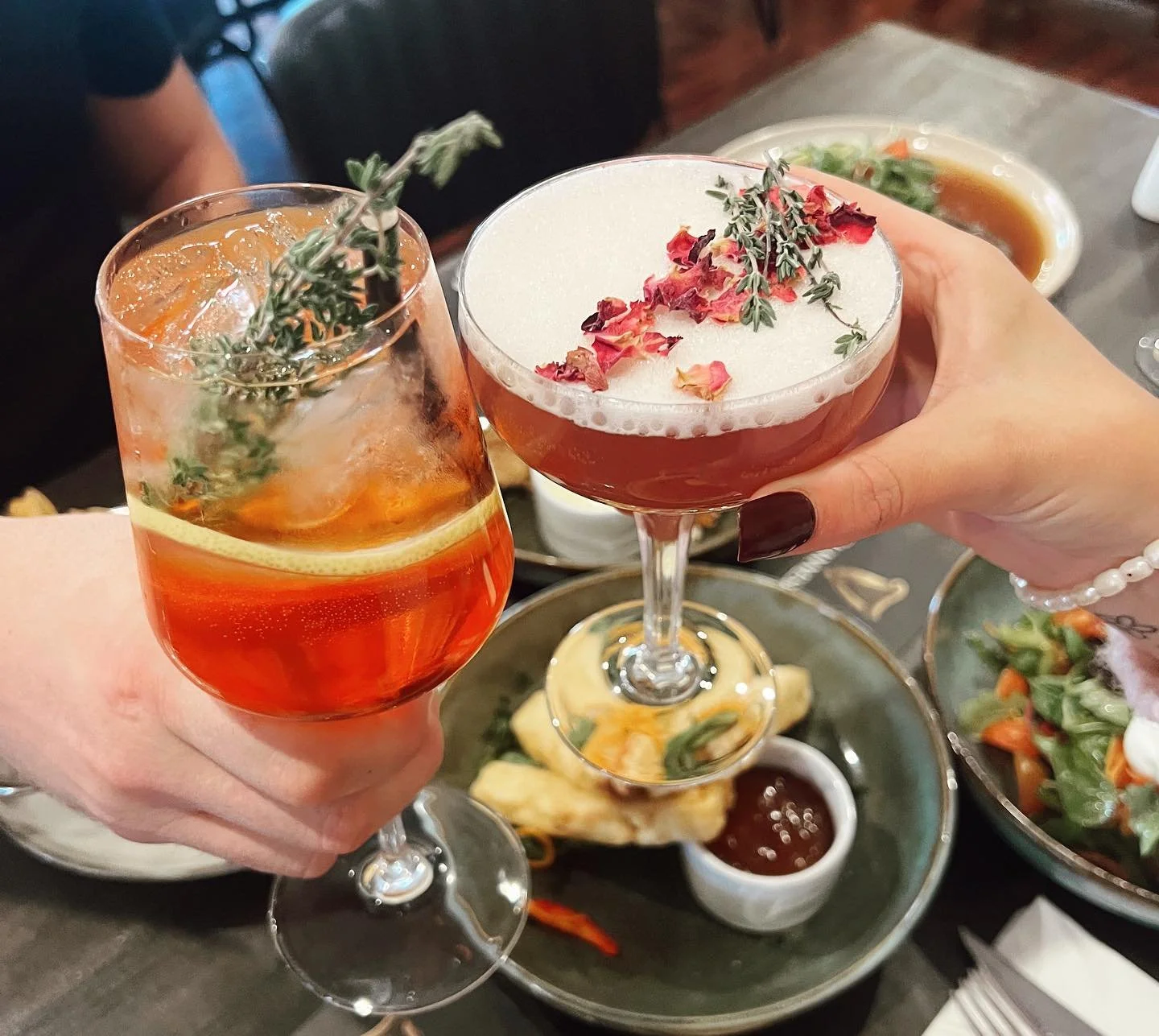 Two people holding cocktails with herbs and flowers, celebrating at a dining table with food dishes in the background.