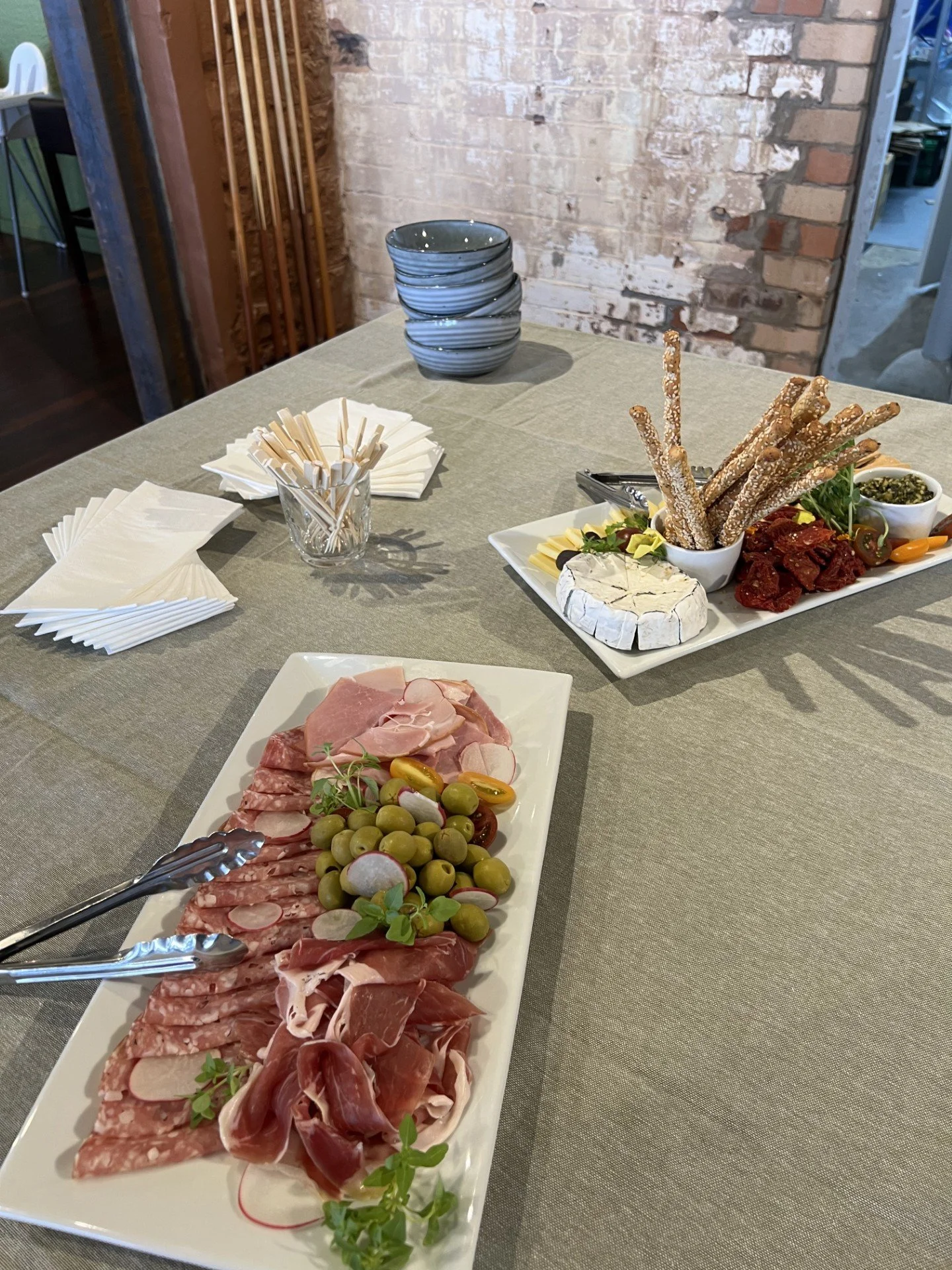 Charcuterie board with cured meats, green olives, and cheese on a table with napkins and a stack of bowls.