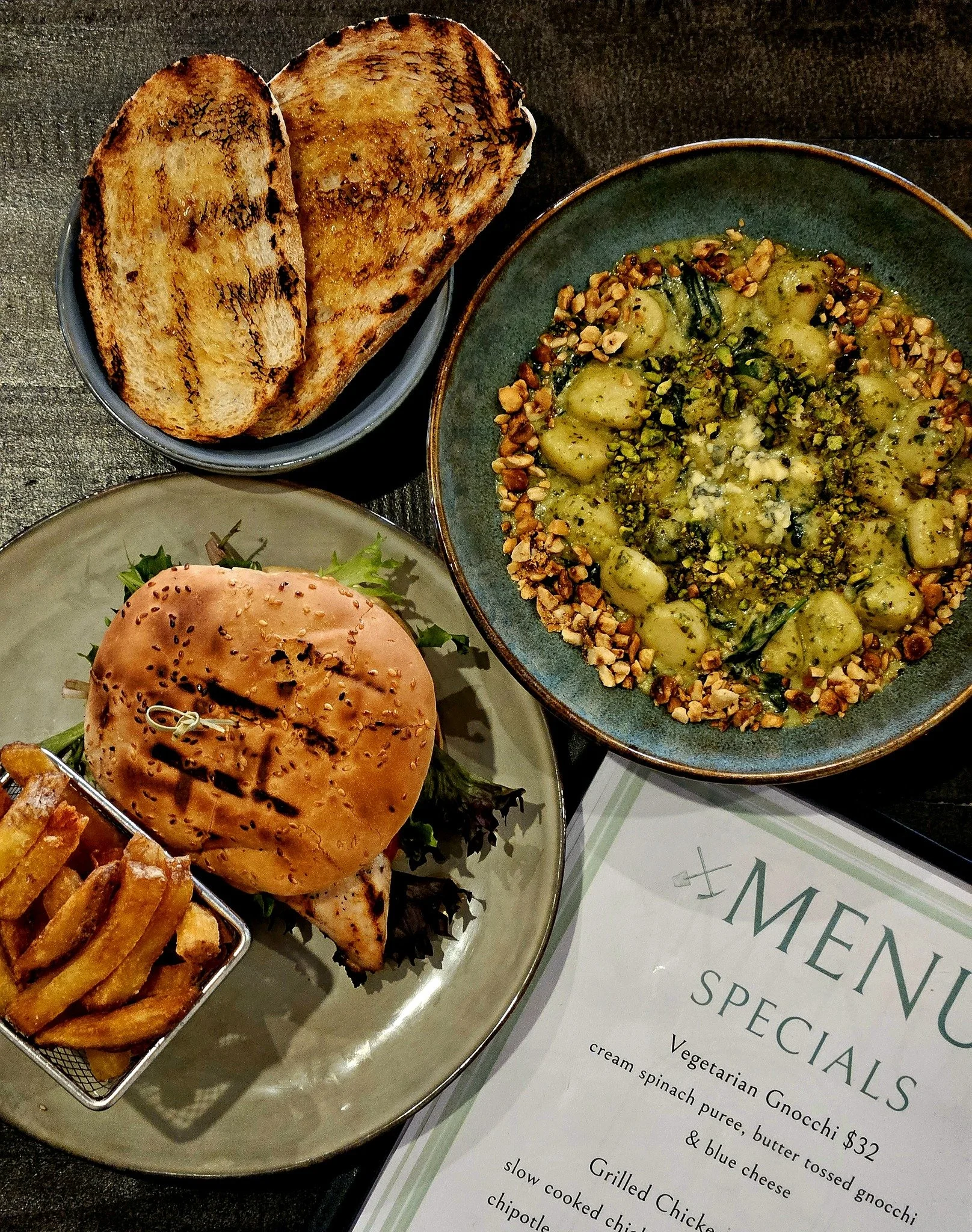 A meal featuring toasted bread, a veggie gnocchi dish with chopped nuts on top, a grilled chicken sandwich with fries, and a menu with vegetarian gnocchi listed.