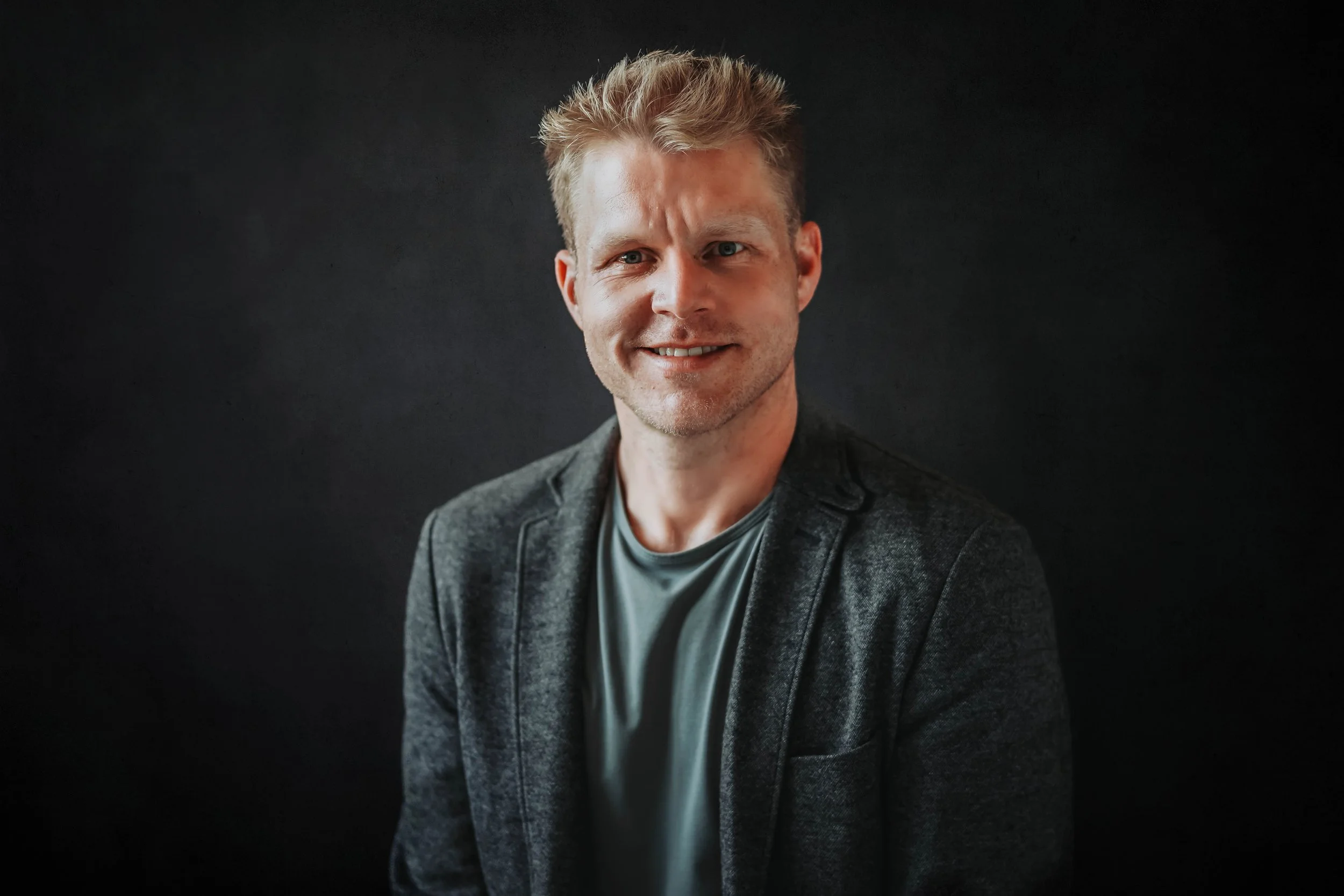 Portrait of a smiling man with light skin, blond hair, wearing a dark blazer and gray shirt against a black background.