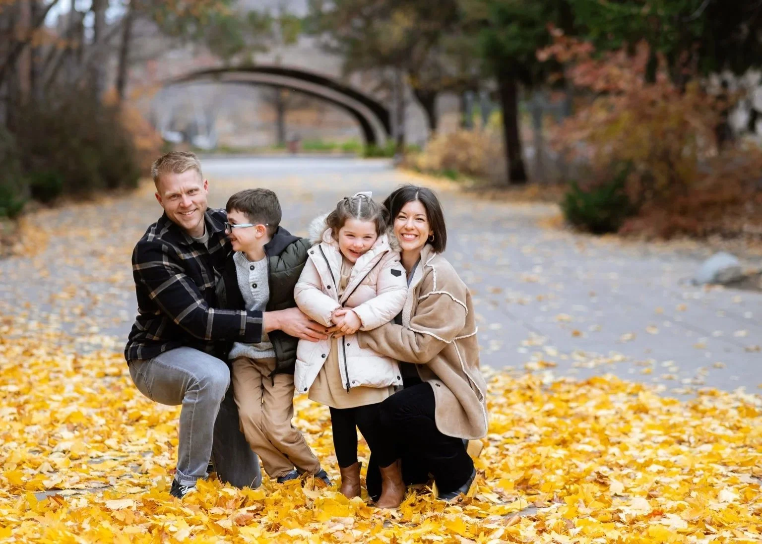 A family of four, including two parents and two children, smiling and hugging on a leaf-covered path in a park during fall.