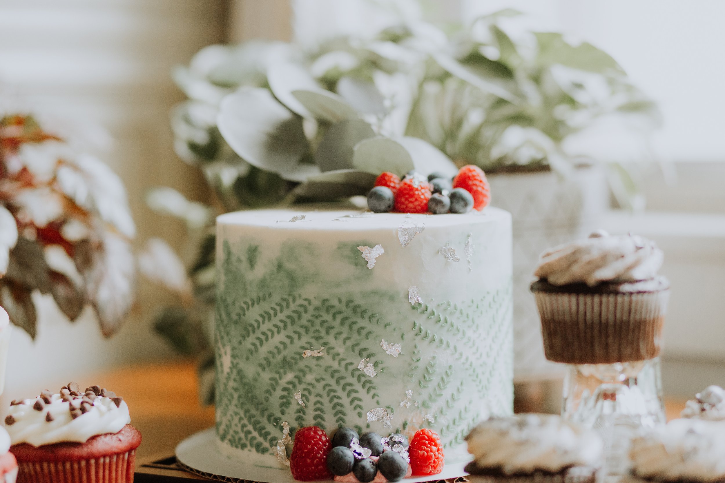 Decorative cake with green icing and berries, surrounded by cupcakes.