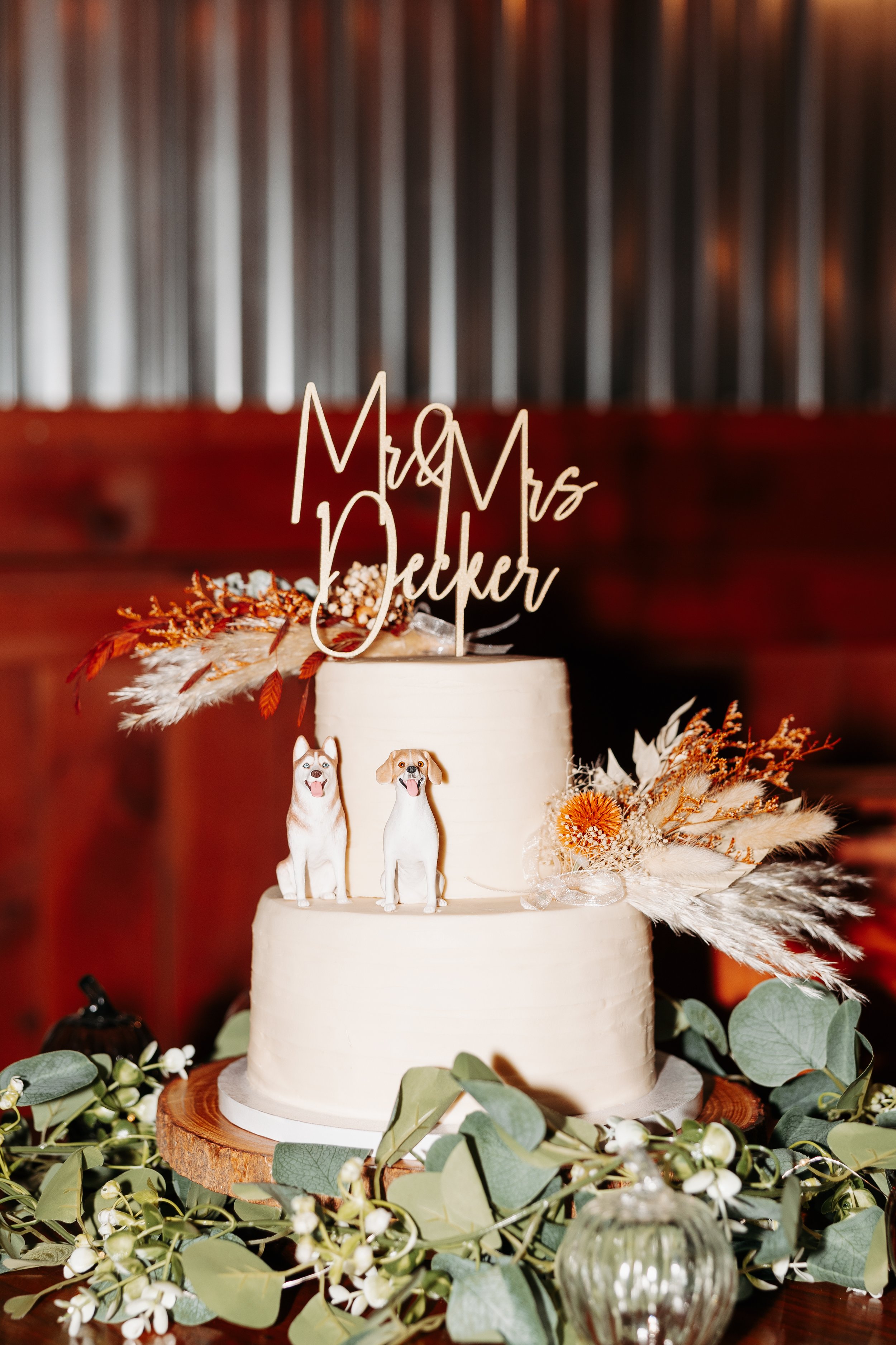 Two-tier white wedding cake with dog figurines, dried flowers, and 'Mr & Mrs Decker' topper, surrounded by greenery on a wood base.