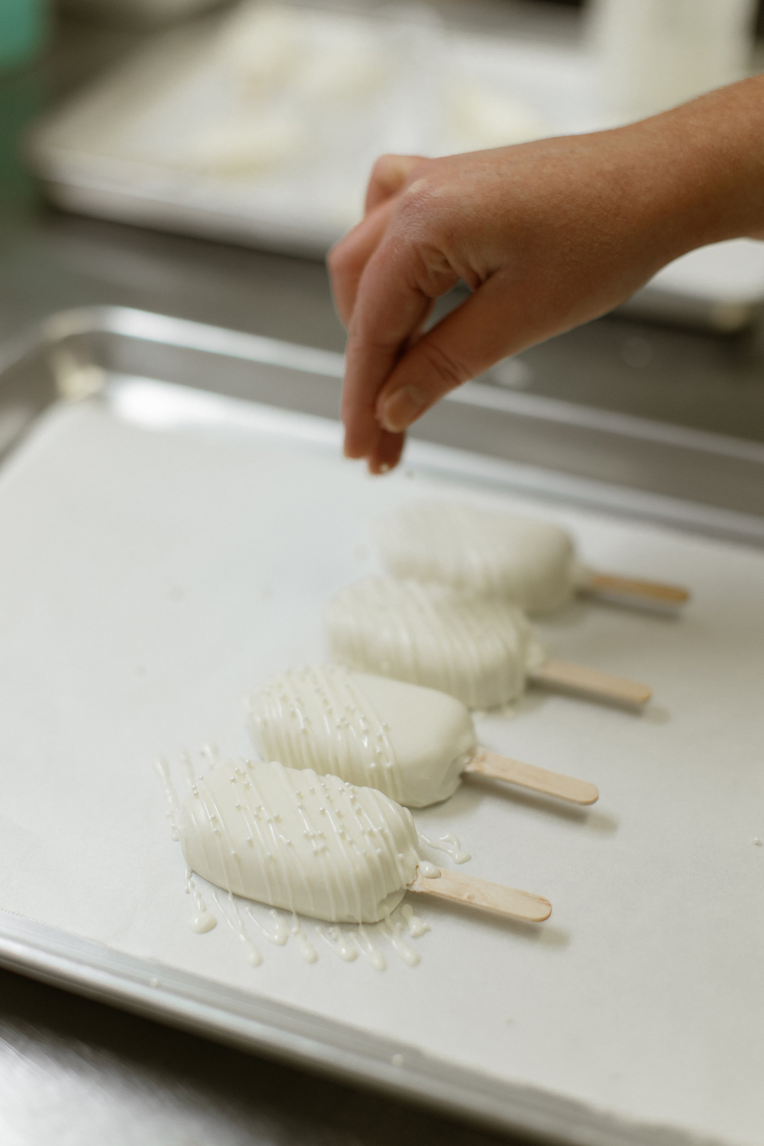 Hand sprinkling toppings on white chocolate-covered ice cream bars on a baking sheet.