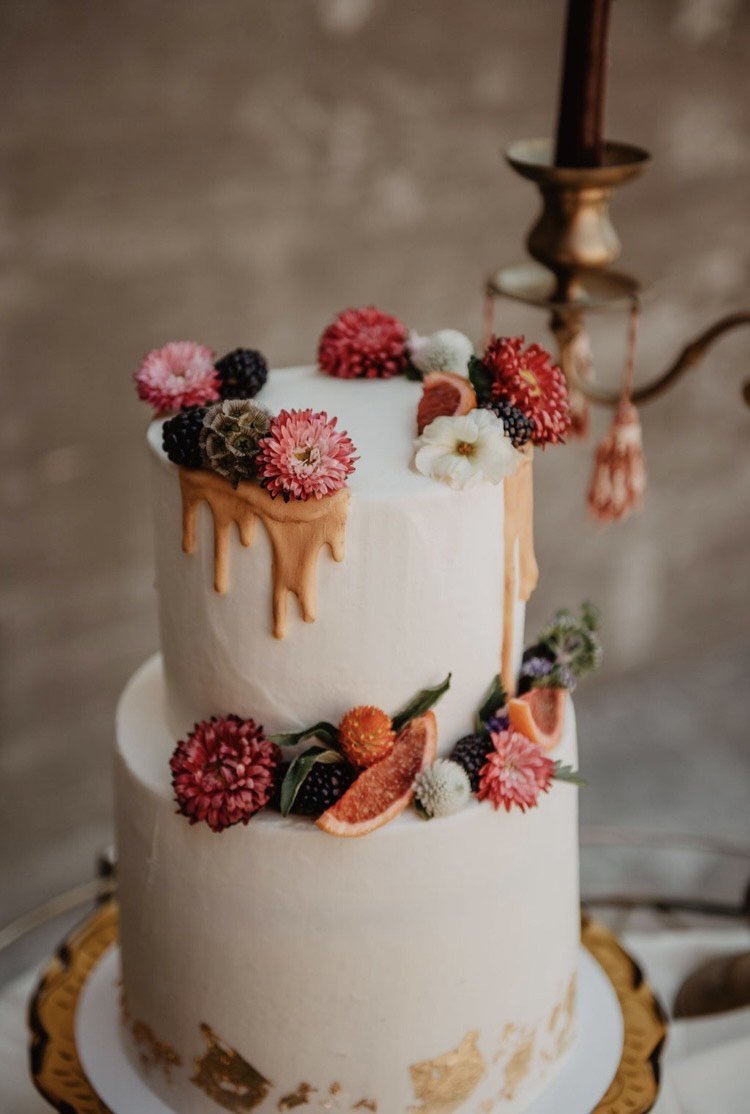 Two-tiered wedding cake with white frosting, gold drip decoration, and adorned with colorful flowers and berries on a gold-trimmed stand.