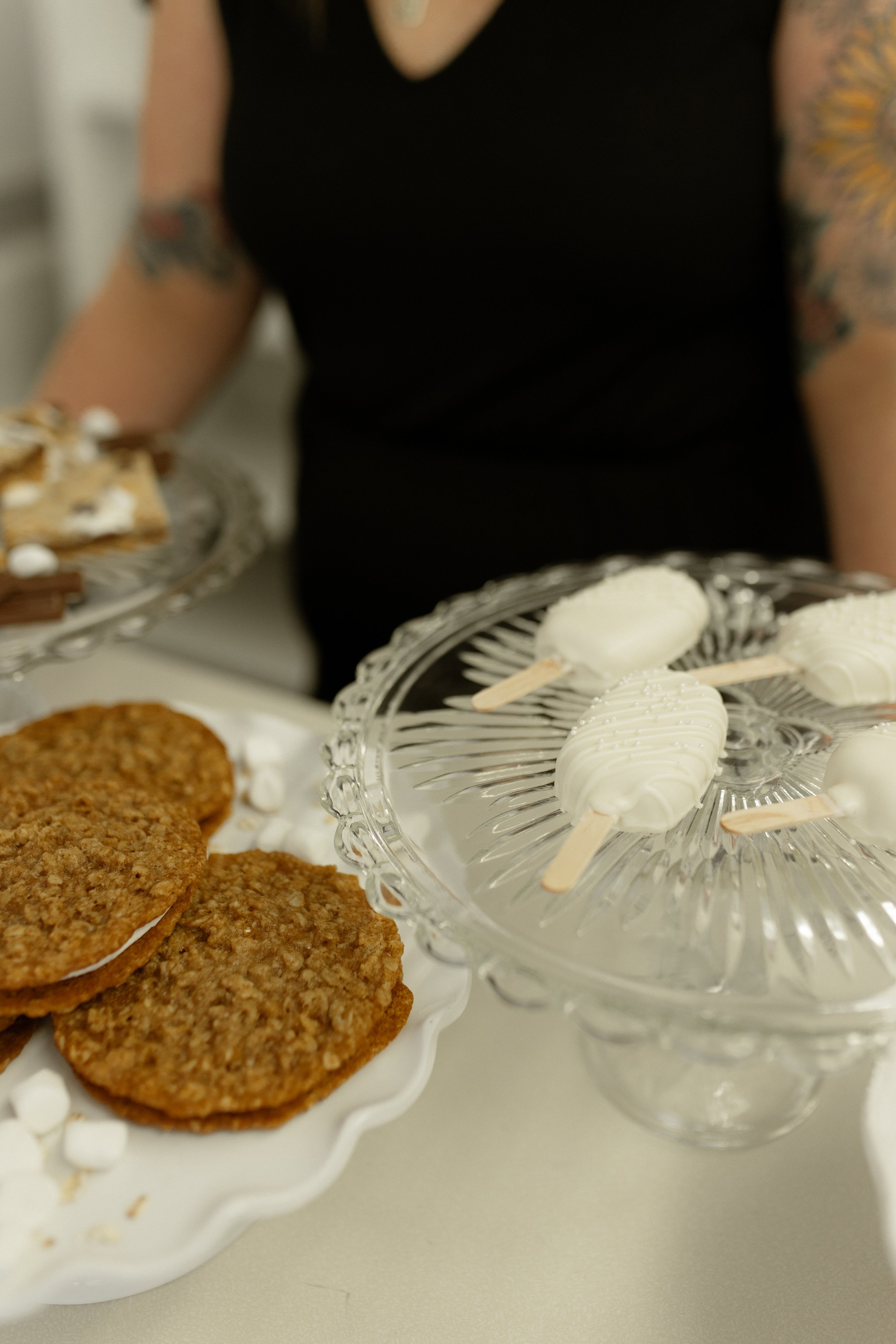 Desserts on glass stands including oatmeal cookies with marshmallow filling and white chocolate-covered treats on sticks. A person in black shirt with tattoos is partially visible.