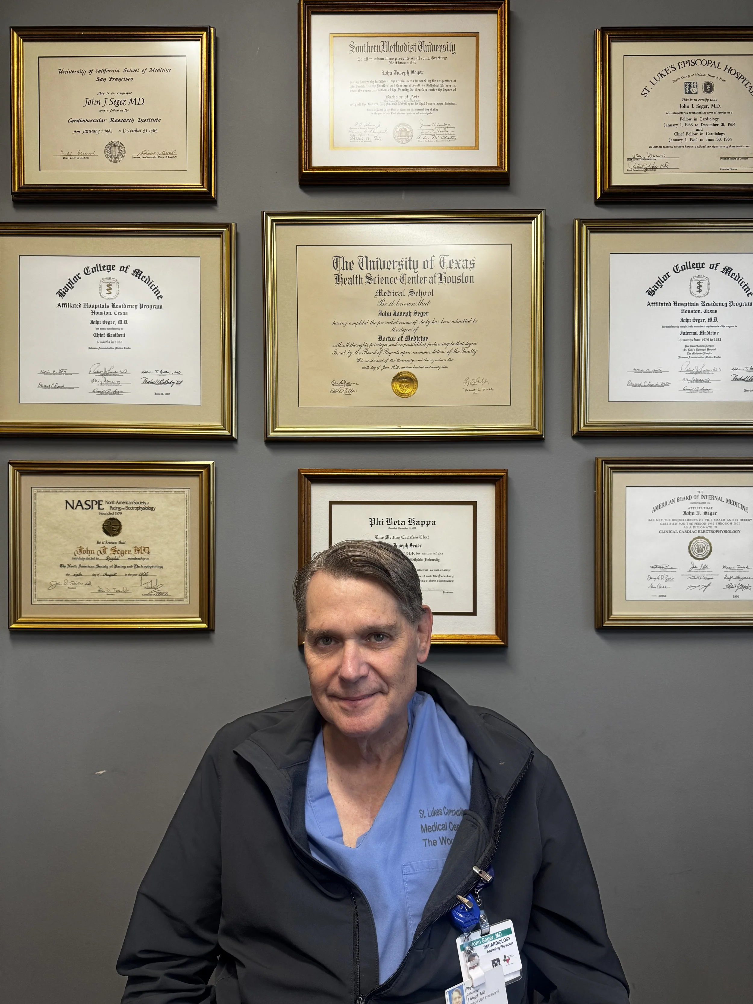 A man in medical scrubs and a black jacket sits in front of a wall with framed diplomas and certificates.
