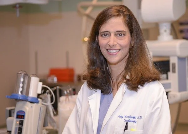 A smiling woman in a white medical coat in a hospital or clinical setting with medical equipment in the background.